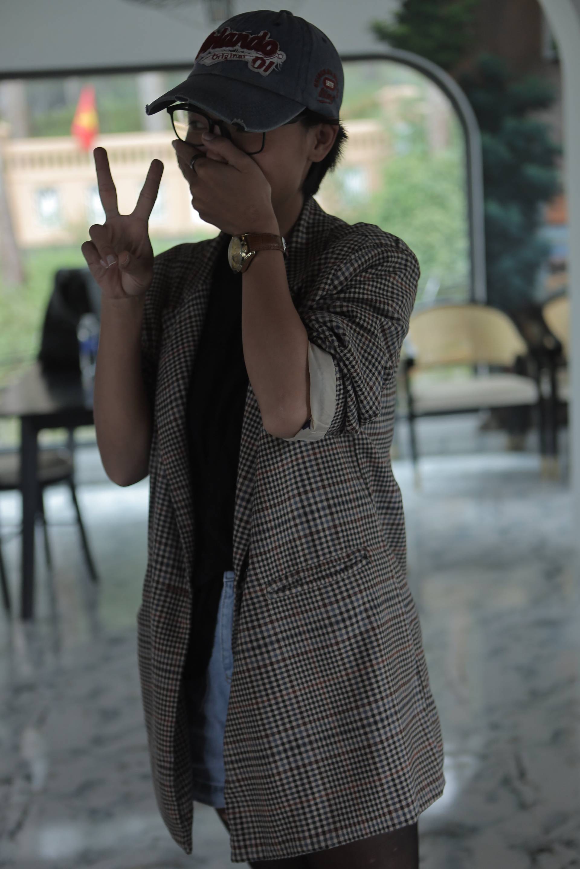 A person in a checkered vest and cap is making a peace sign while standing indoors with a scenic view of Đà Lạt's natural beauty in the background.