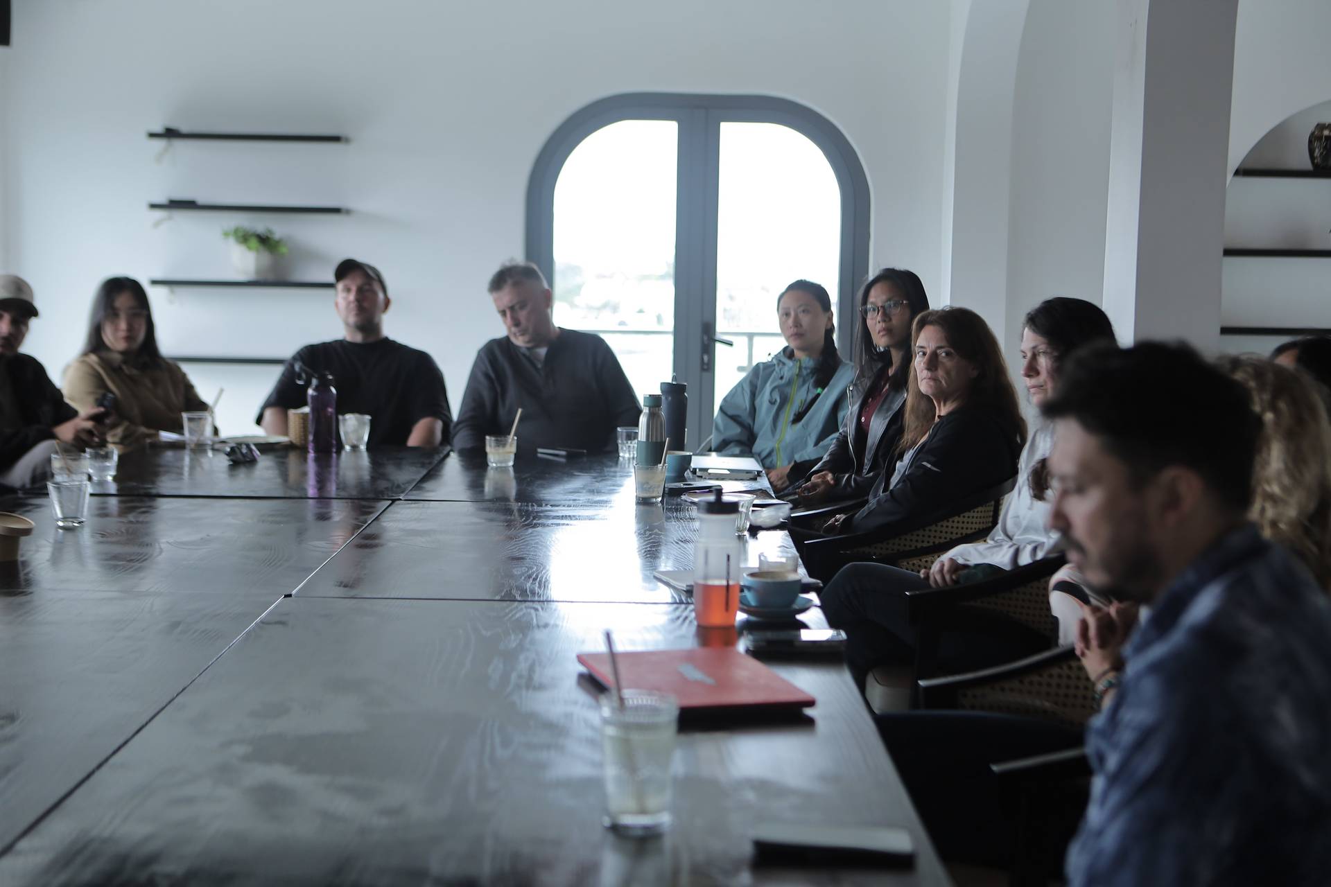 A group of people is seated around a long wooden table in a modern, well-lit room with large windows overlooking Đà Lạt's scenic landscape. They appear to be engaged in a meeting or discussion, with drinks and notebooks on the table.