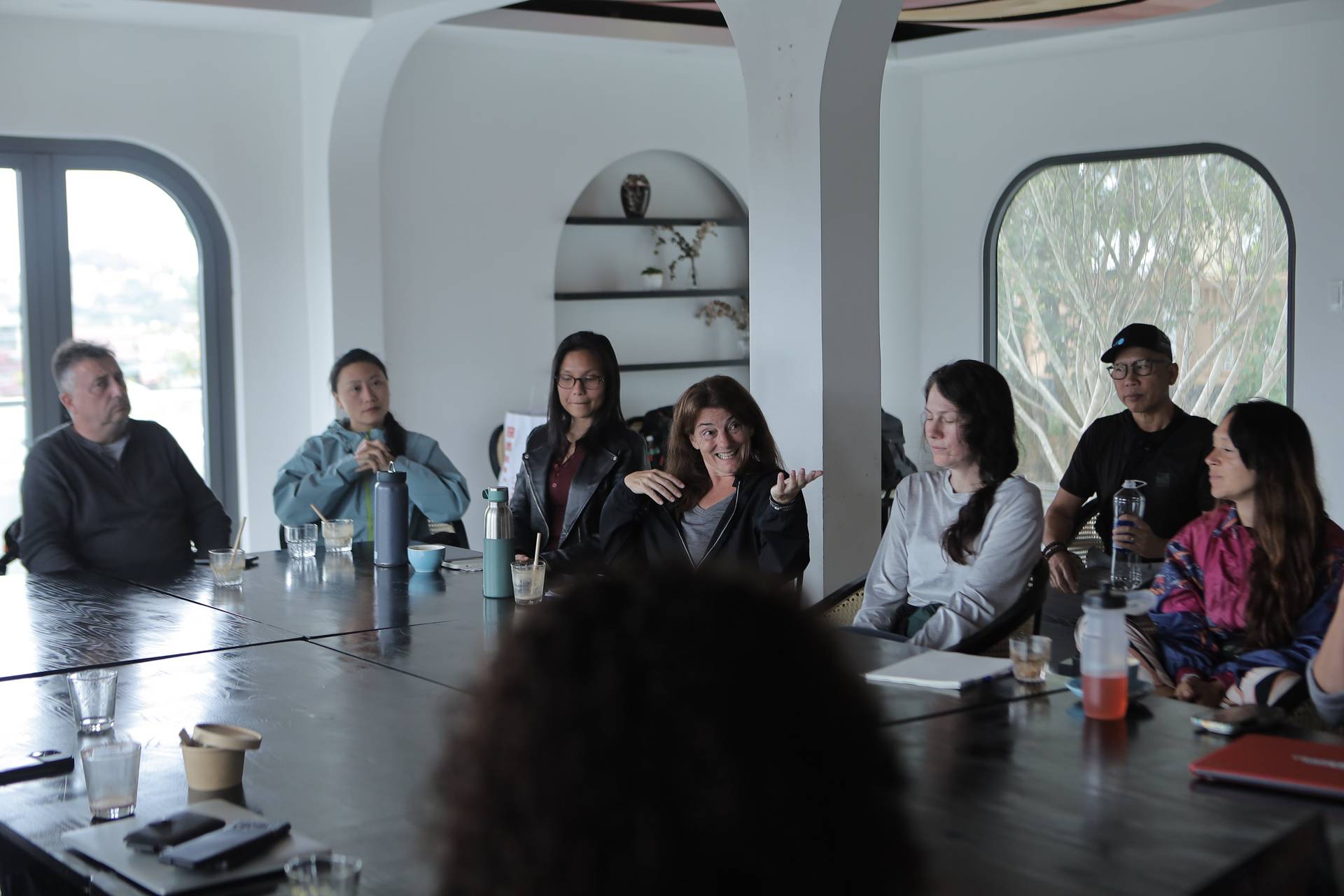 A group of people is gathered around a long table in a modern, well-lit room with large windows overlooking an urban landscape. They appear to be engaged in a casual meeting or discussion, enjoying drinks and snacks.