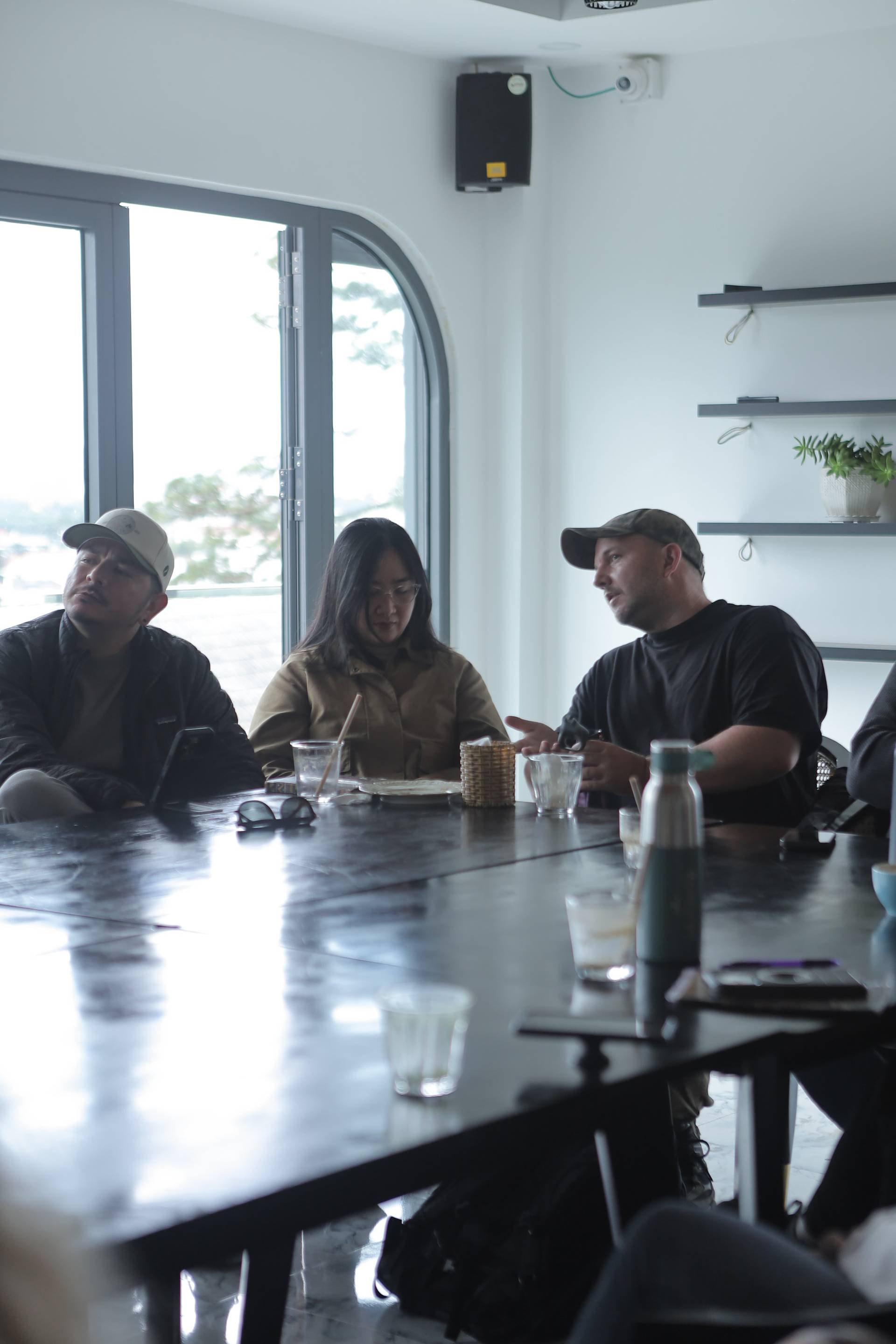 A group of individuals engaged in a casual meeting or discussion around a table in a modern café setting with large windows overlooking a serene outdoor view.