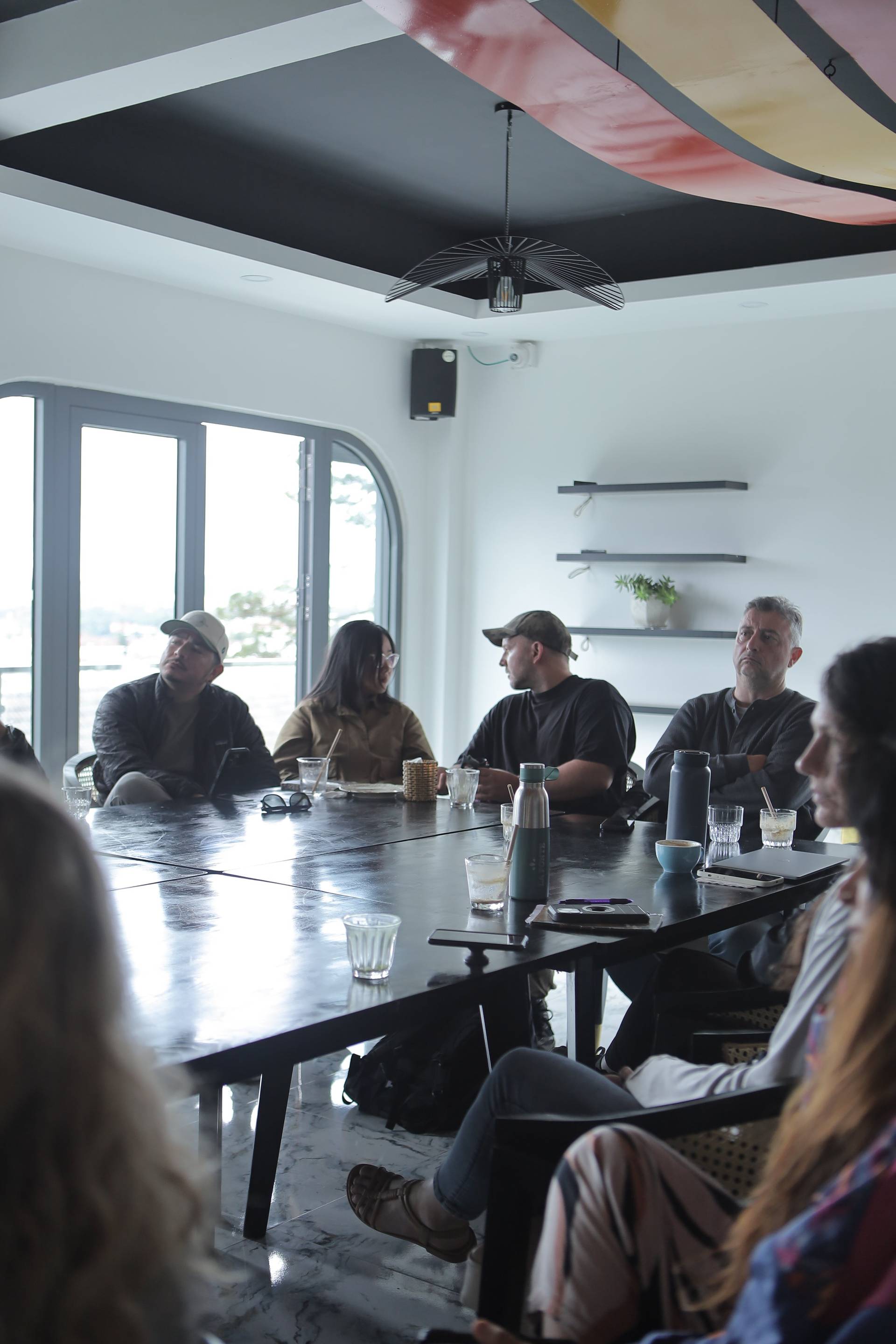 A group of people is gathered around a long table in a modern café setting in Đà Lạt, Vietnam, engaged in conversation and enjoying drinks.