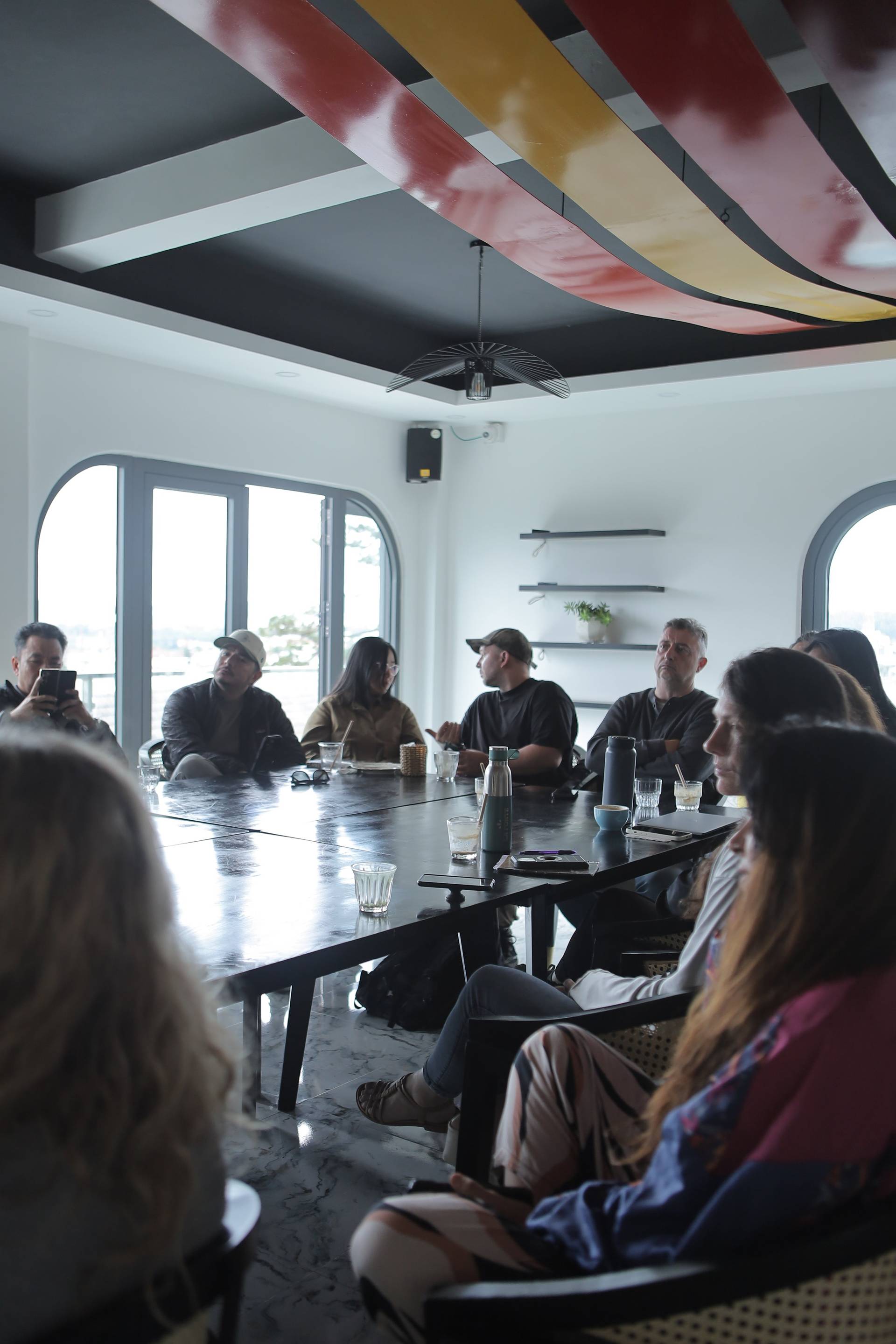 A group of people is gathered around a long table in a modern café with large windows overlooking Đà Lạt's scenic landscape, engaging in conversation and enjoying drinks.