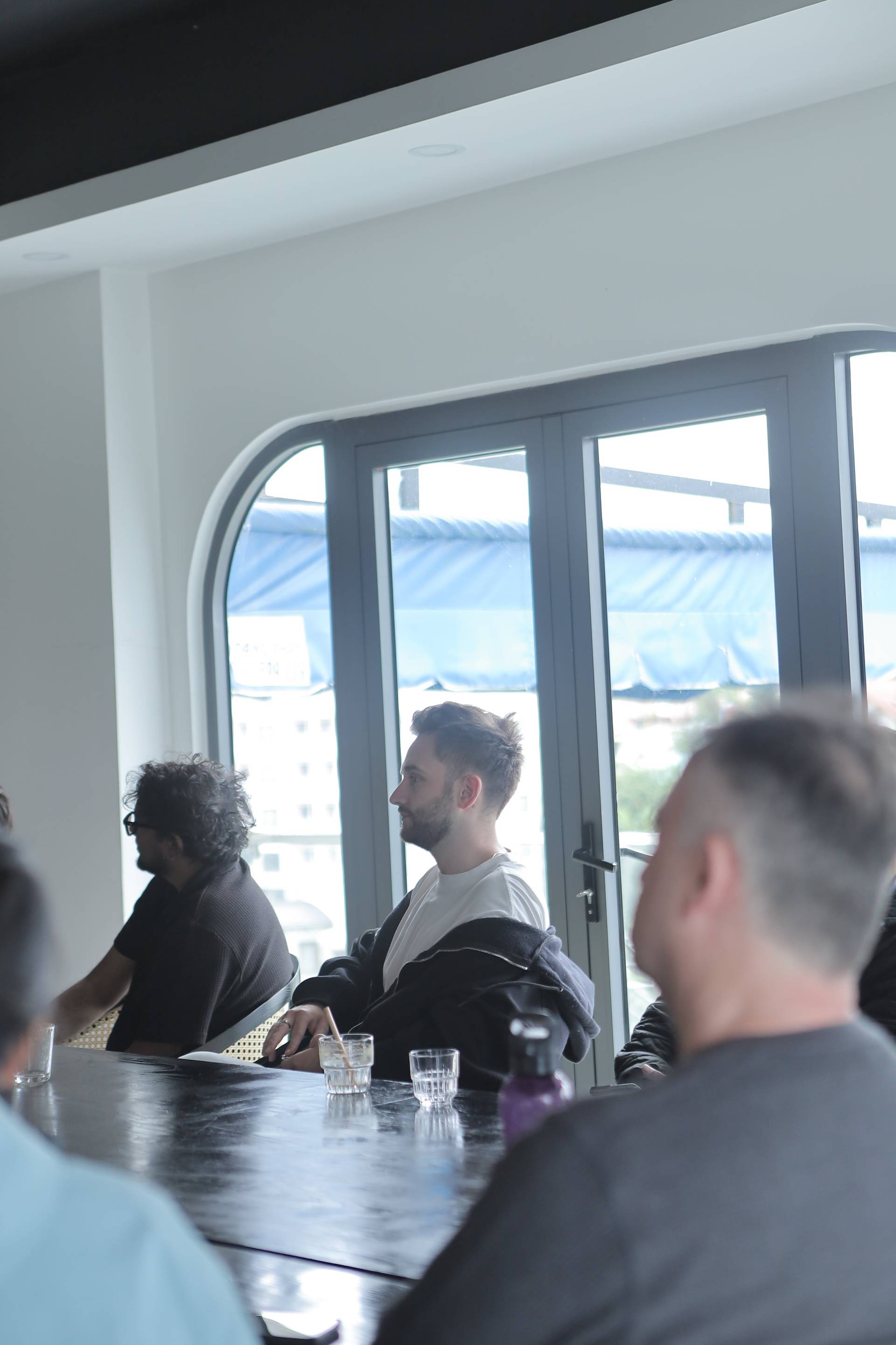 A group of individuals is gathered around a table in a modern indoor setting with large windows overlooking a cityscape, engaging in conversation and enjoying drinks.