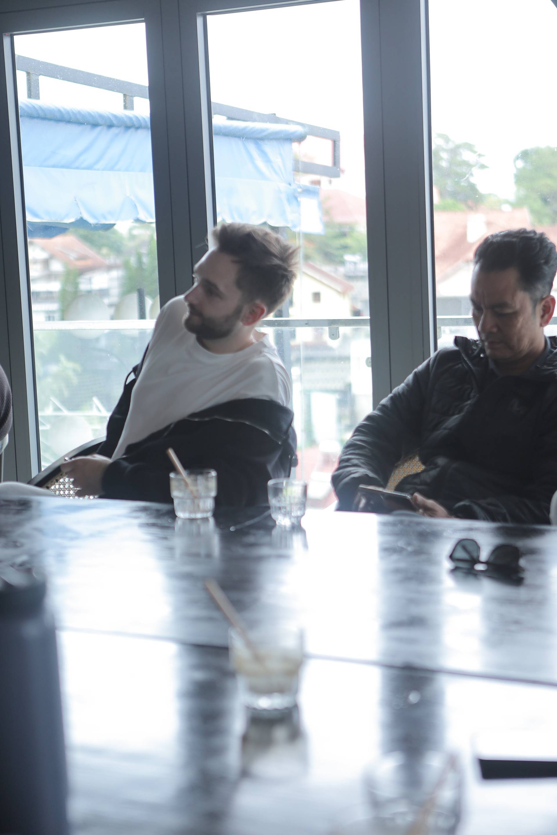 Two individuals engage in a casual conversation over drinks by large windows overlooking a serene neighborhood in Đà Lạt, Vietnam.
