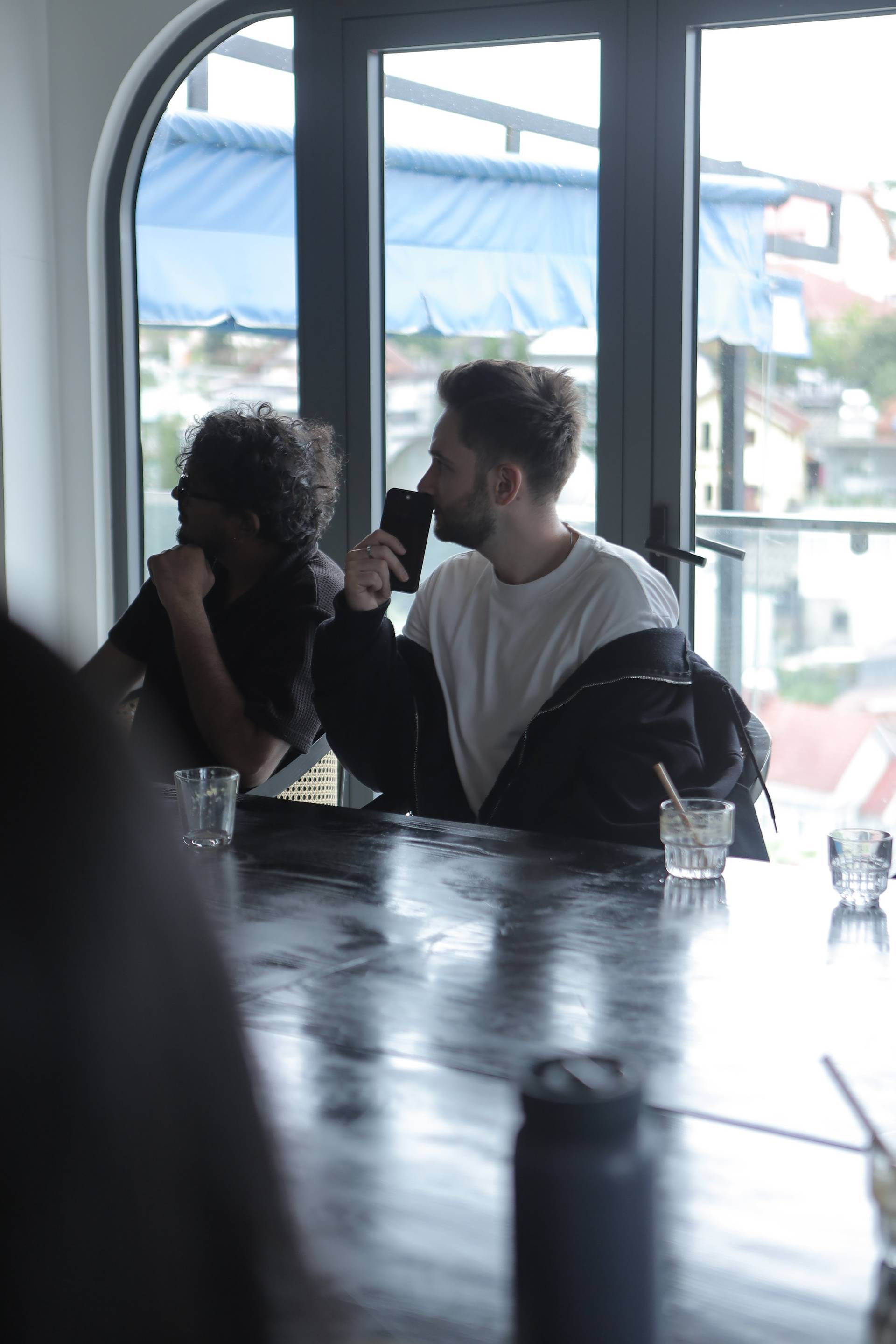 Two individuals engage in a quiet conversation over drinks by a large window overlooking Đà Lạt's scenic landscape.