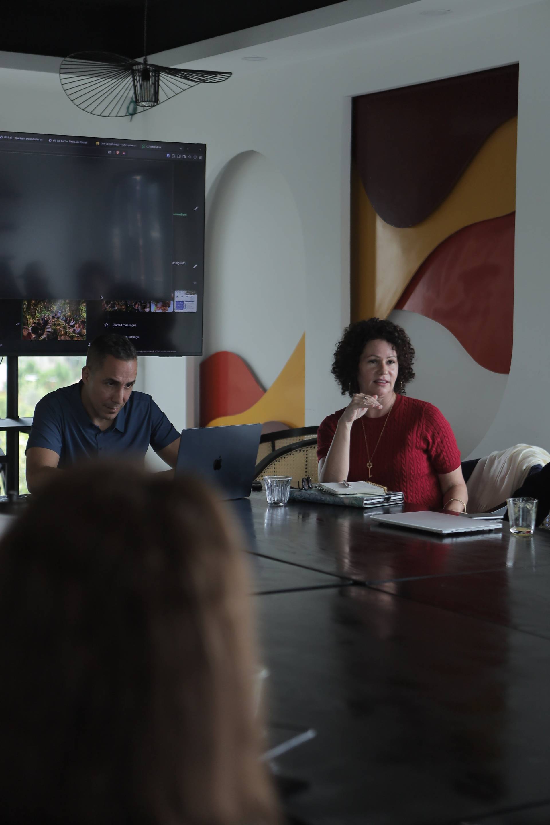 A professional meeting in a modern conference room with attendees engaged in discussion, featuring a large screen displaying content and colorful abstract wall art.