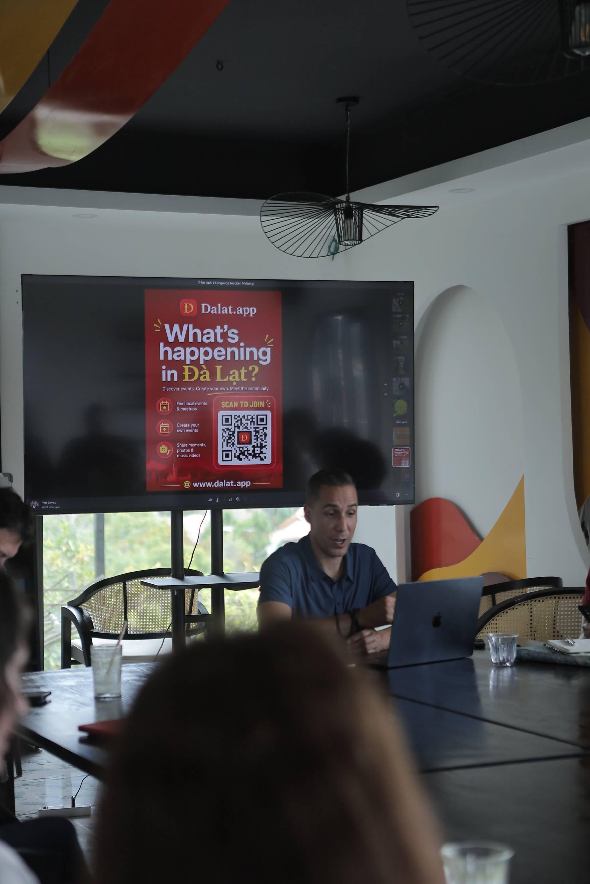 A presentation about local events in Đà Lạt is being held in a modern café with attendees seated around a table, engaging with the content displayed on a large screen.