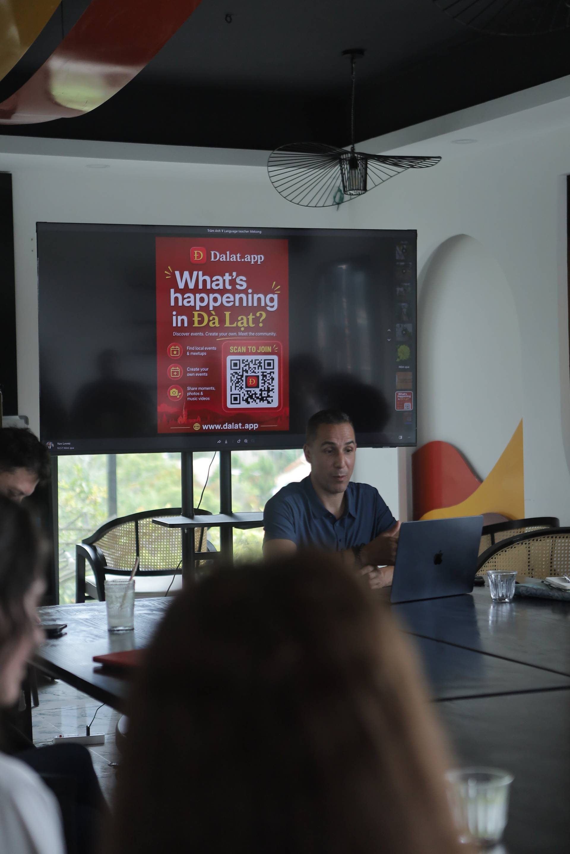 A presentation about events in Đà Lạt is being given to a small audience in a modern meeting room with a large screen displaying promotional content for Dalat.app.