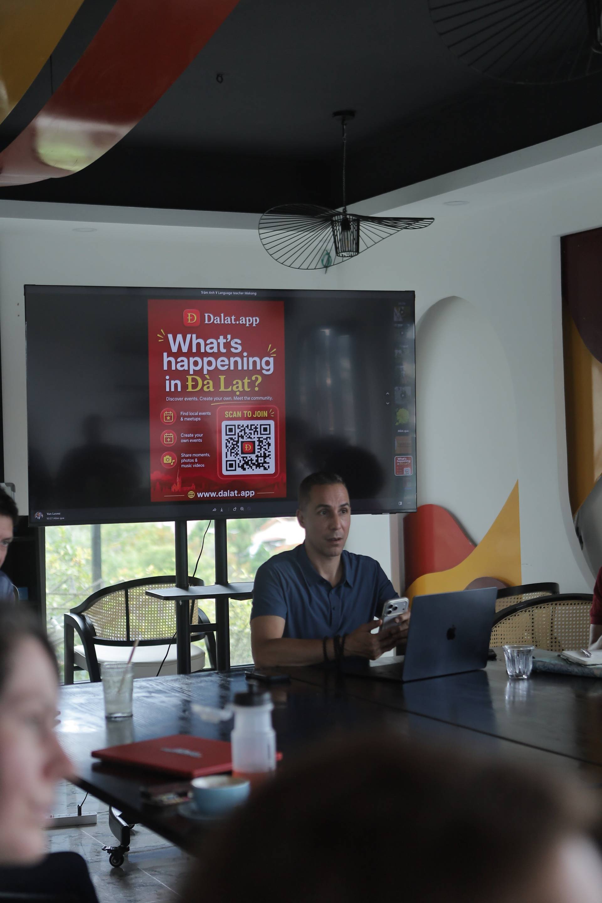 A presentation about local events in Đà Lạt is being given to a small group of attendees seated around a table with modern decor and a large screen displaying promotional content for Dalat.app.