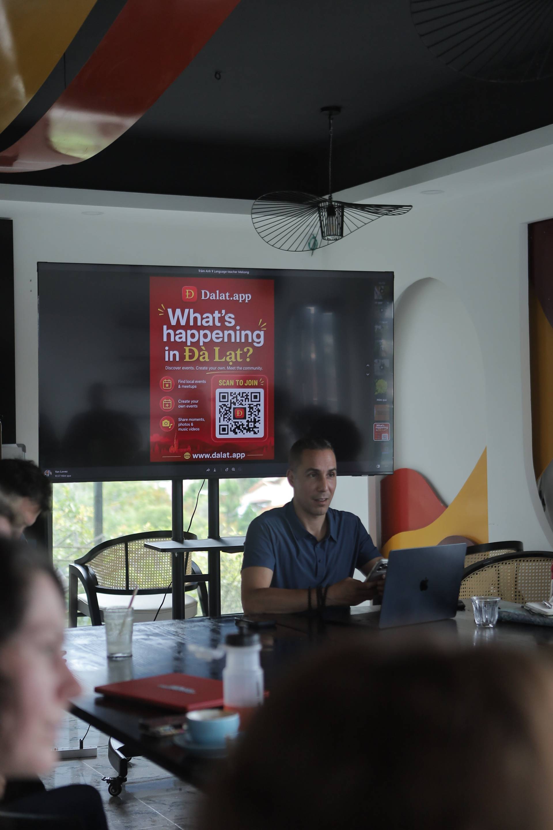 A presentation about events in Đà Lạt is being held in a modern café with attendees seated around a table, engaging with content displayed on a large screen.
