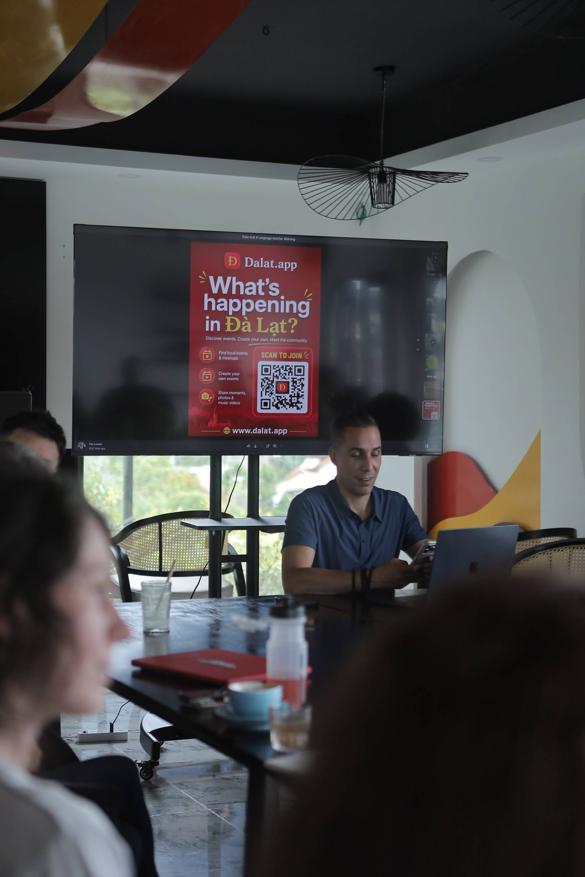 A group of people is gathered in a modern café in Đà Lạt, Vietnam, engaging with a presentation about local events and community engagement using Dalat.app.