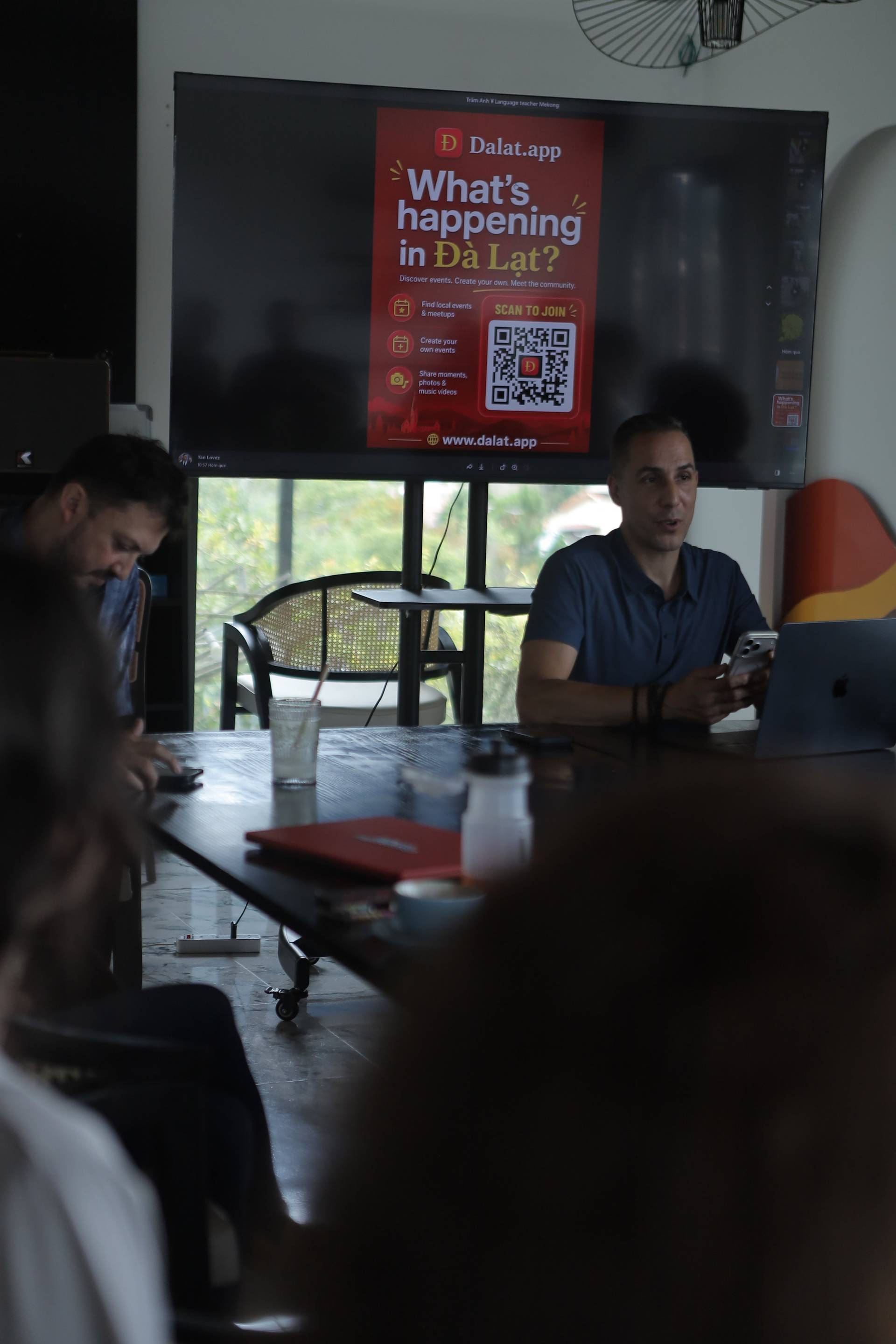 A group of people is gathered around a table in Đà Lạt, Vietnam, discussing and using a digital app to explore local events and community activities.