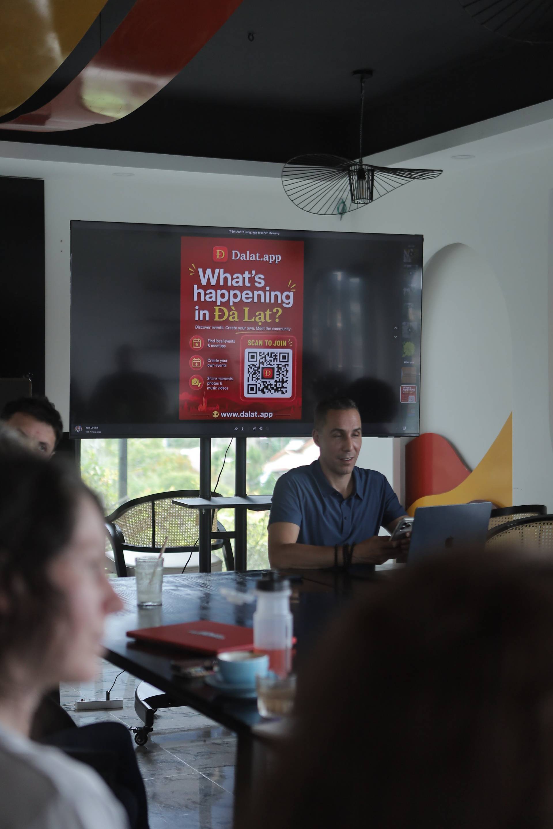 A presentation about Dalat.app is being given in a modern café setting with attendees seated around a table, engaging with the content displayed on a large screen.