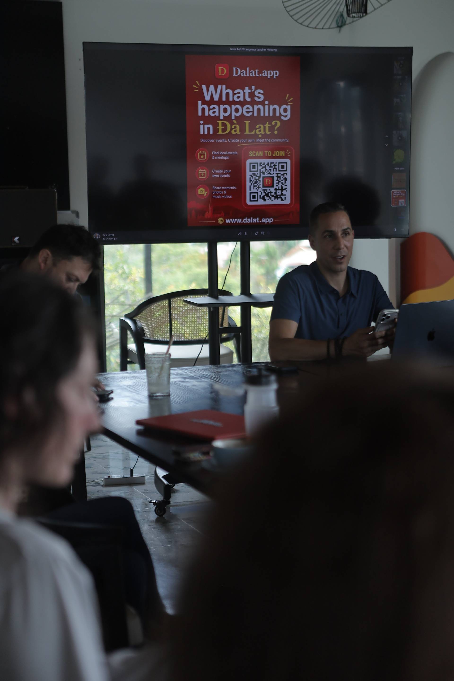 A group of people is gathered in a cozy café in Đà Lạt, Vietnam, discussing and presenting about the local app 'Dalat.app', which promotes events and community engagement.