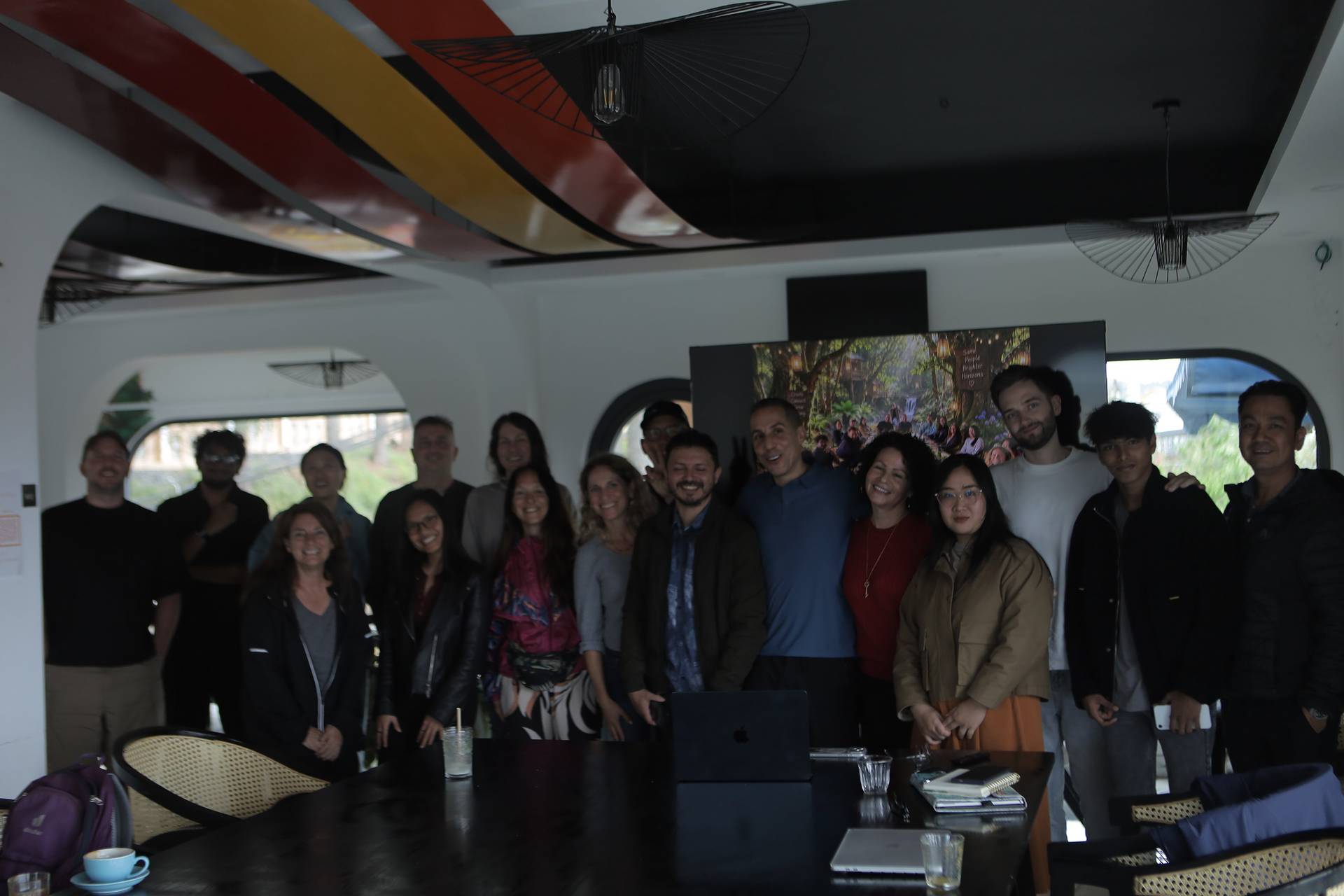 A group of people is gathered in a modern, vibrant space with colorful ceiling art and large windows overlooking greenery, smiling and posing together for a photo.