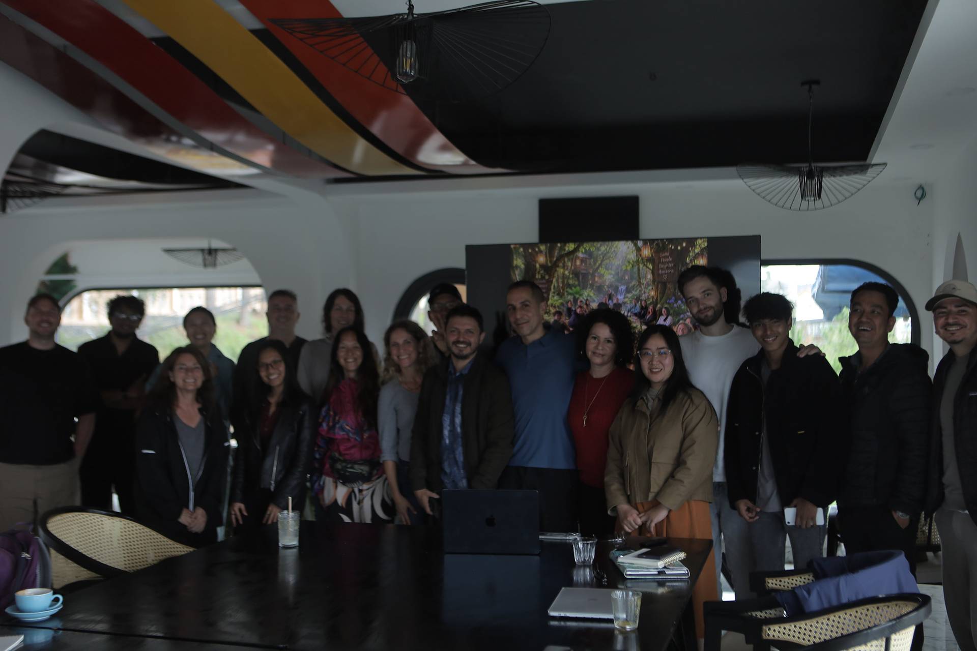 A group of people gathered in a modern, stylish room with artistic ceiling decorations and a scenic poster in the background, smiling and posing together for a photo.