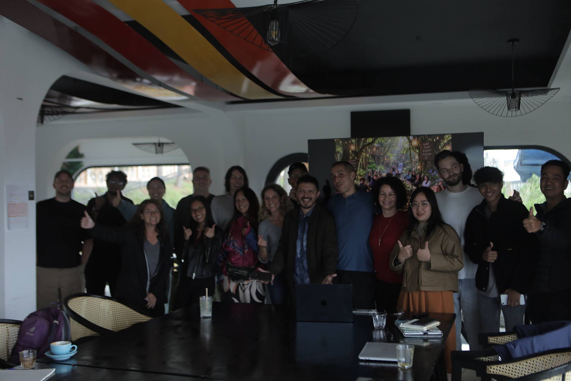 A group of people is gathered in a modern, stylish room with vibrant decor and large windows overlooking Đà Lạt's scenic landscape, smiling and giving thumbs up for a cheerful team photo.