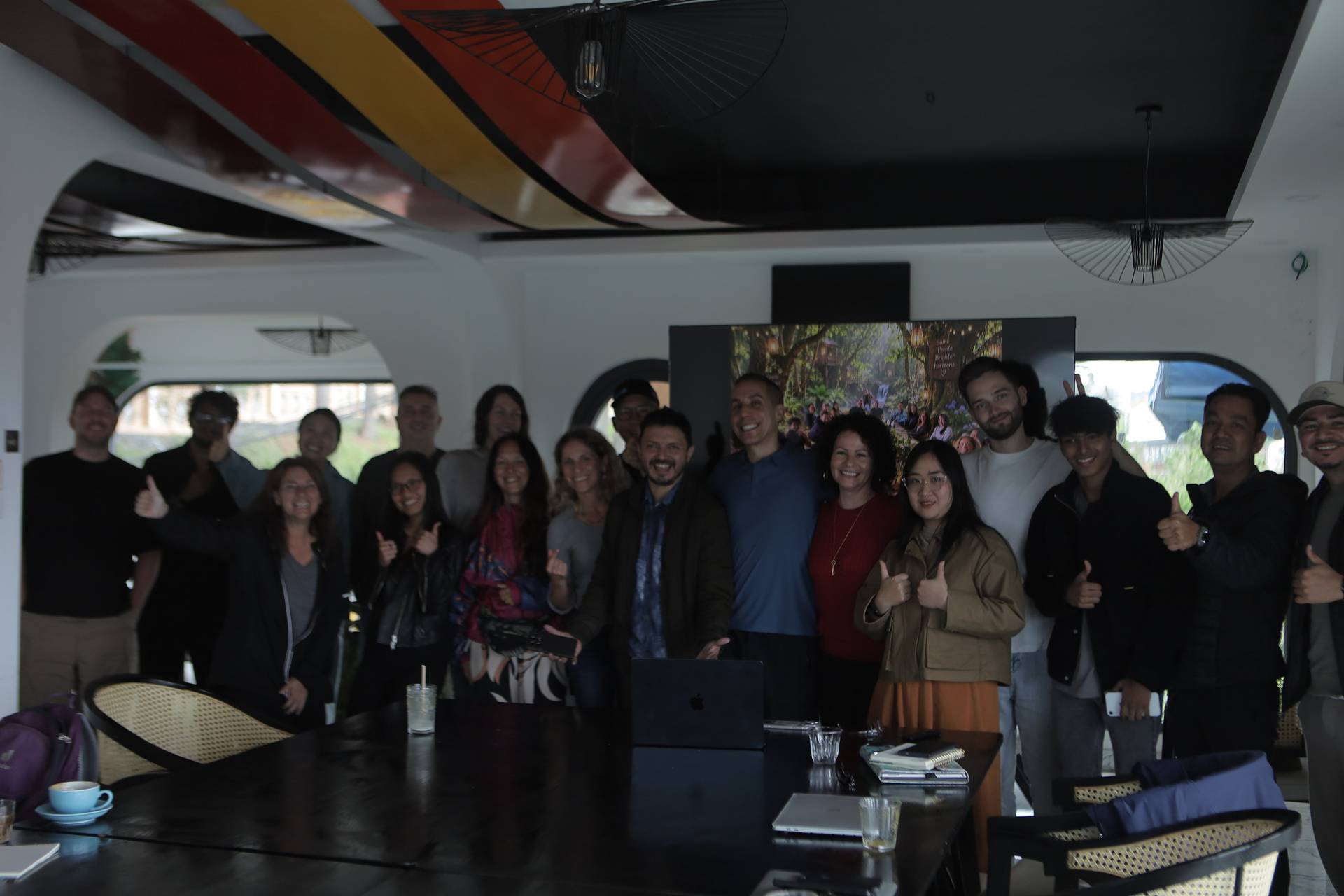 A group of people is gathered in a modern, well-lit room with artistic decor in Đà Lạt, Vietnam, smiling and giving thumbs up, suggesting a positive and celebratory mood.