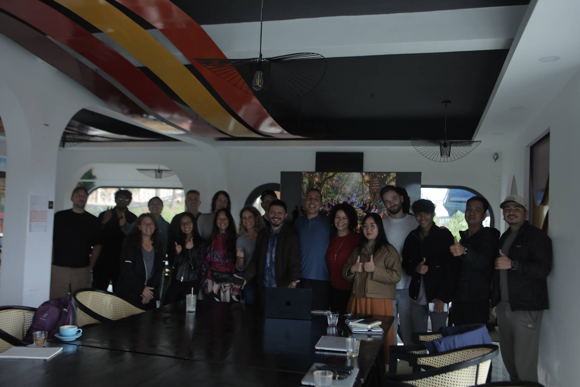 A group of people is gathered in a modern office space with vibrant ceiling art and large windows overlooking greenery, smiling and posing together for a photo.