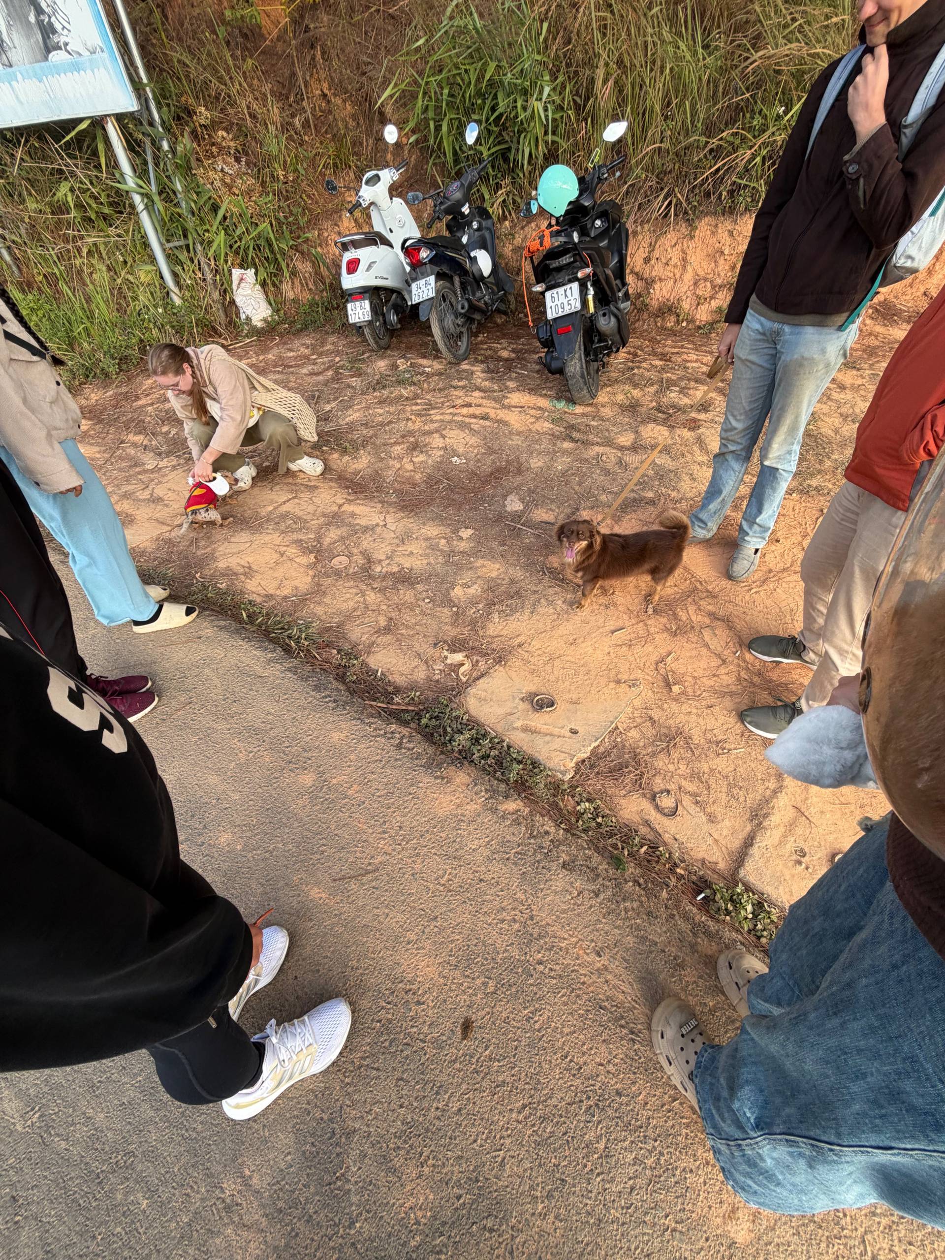 A group gathers around motorcycles on a rural forest path in Đà Lạt during an outdoor motorbike touring adventure. The red-earth setting captures the casual camaraderie of exploring the highlands by scooter.