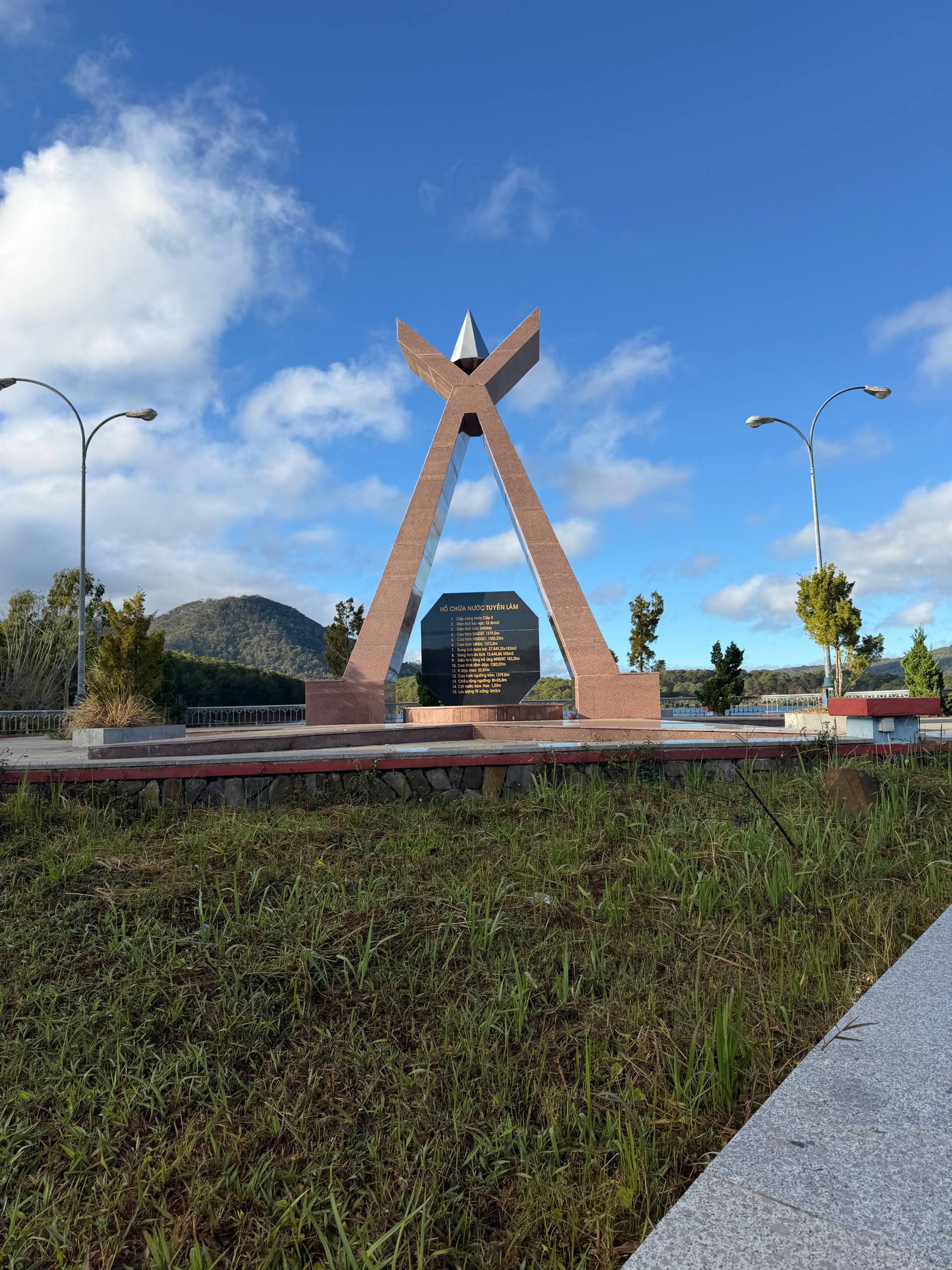 Memorial monument with distinctive X-shaped brick structure and commemorative plaque in landscape setting. Historic landmark in Đà Lạt region with forested hills backdrop.