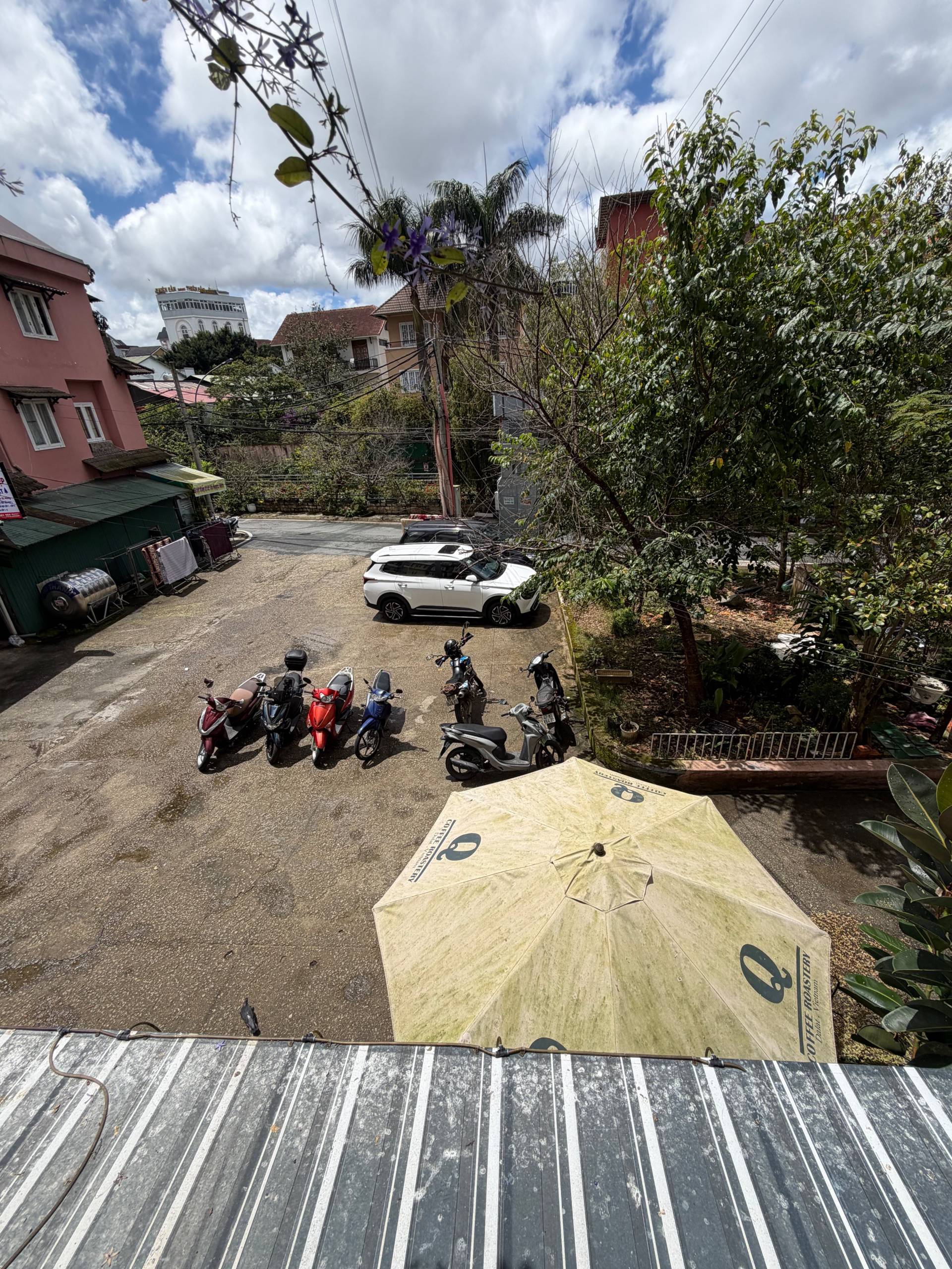 A serene street scene in Đà Lạt, Vietnam, featuring parked scooters and a white car under a cloudy sky with lush greenery surrounding the area.