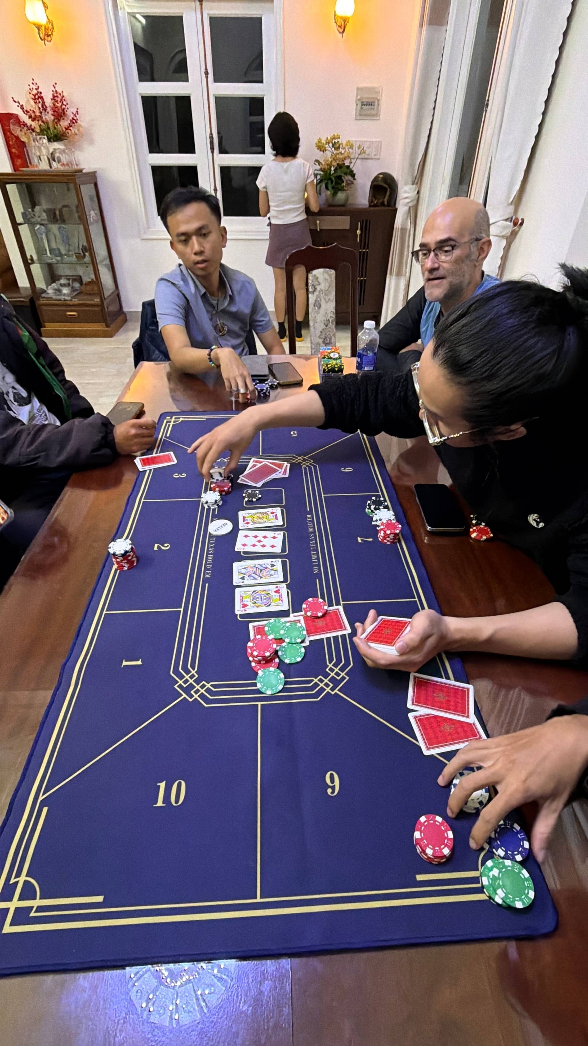 Players place strategic bets on a blue baccarat mat as cards are dealt and colorful chips are arranged in betting patterns. The dynamic scene captures the tension and decision-making moments of an engaging card game in a warmly-lit Vietnamese setting.