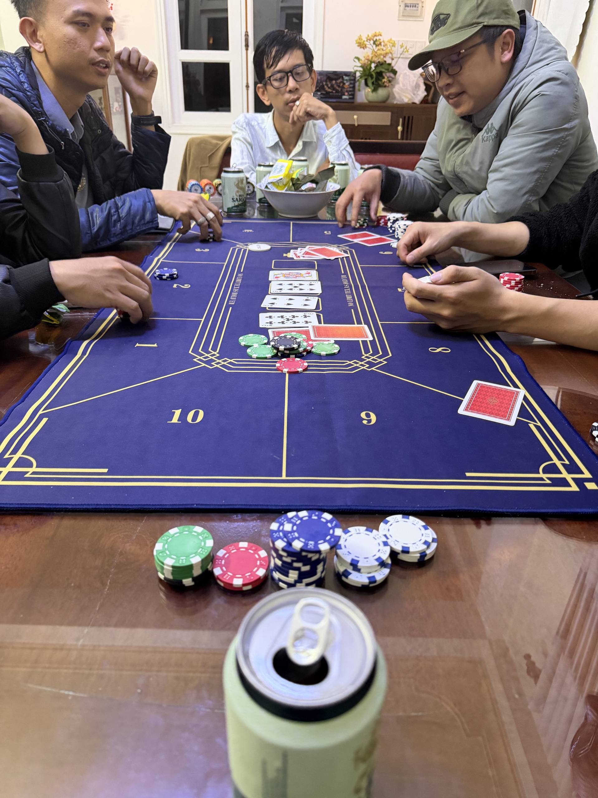 A sophisticated group plays baccarat in an elegantly appointed room featuring large windows, cream drapery, and vintage furnishings. The refined setting combines Vietnamese hospitality with classic gaming traditions, creating an atmosphere of leisure and social connection.