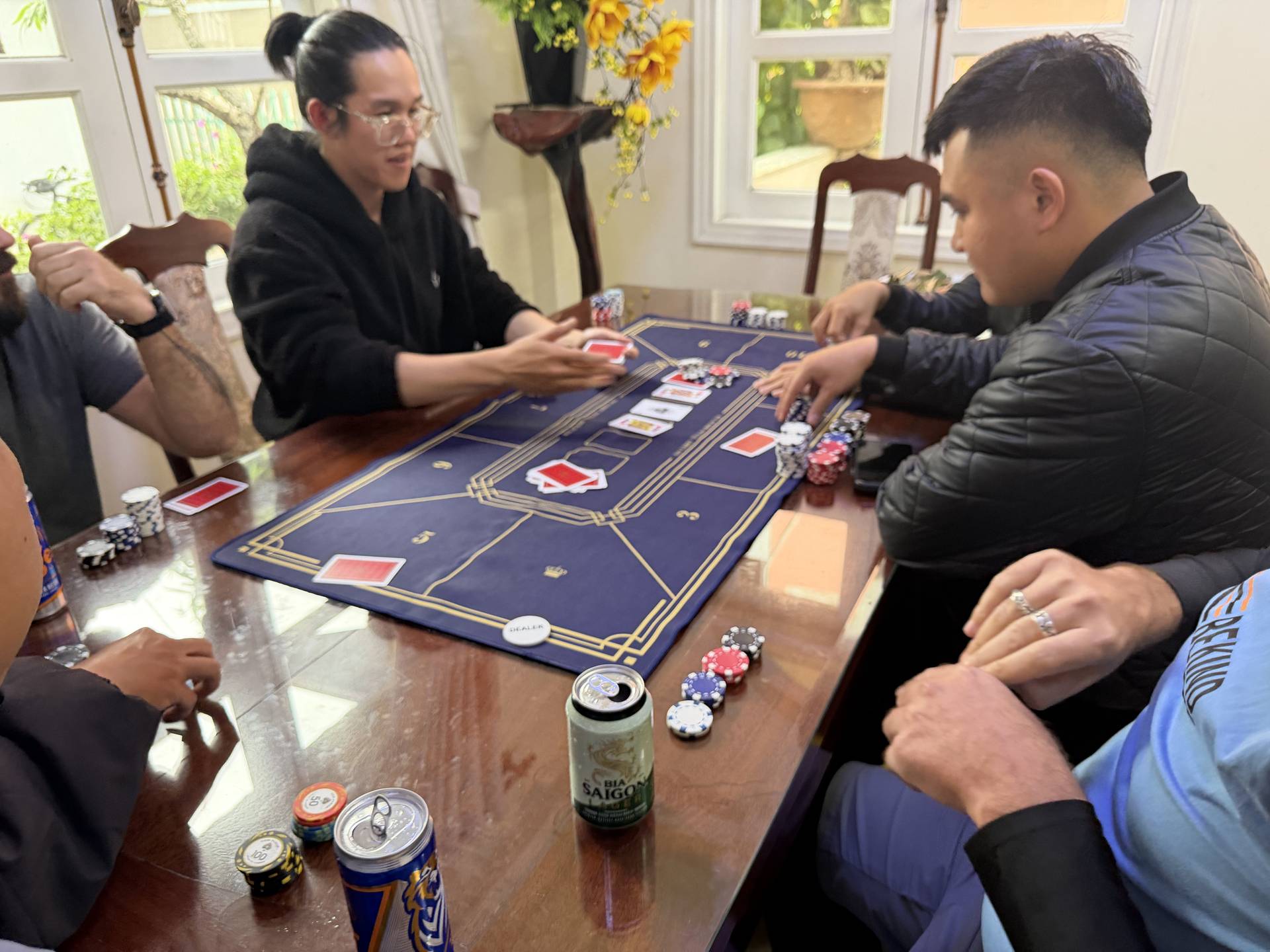 Hands hover over a blue gaming mat as multiple players place bets and manage cards in a casual baccarat session. Colorful poker chips and playing cards capture the excitement of a social card game in an intimate indoor setting.