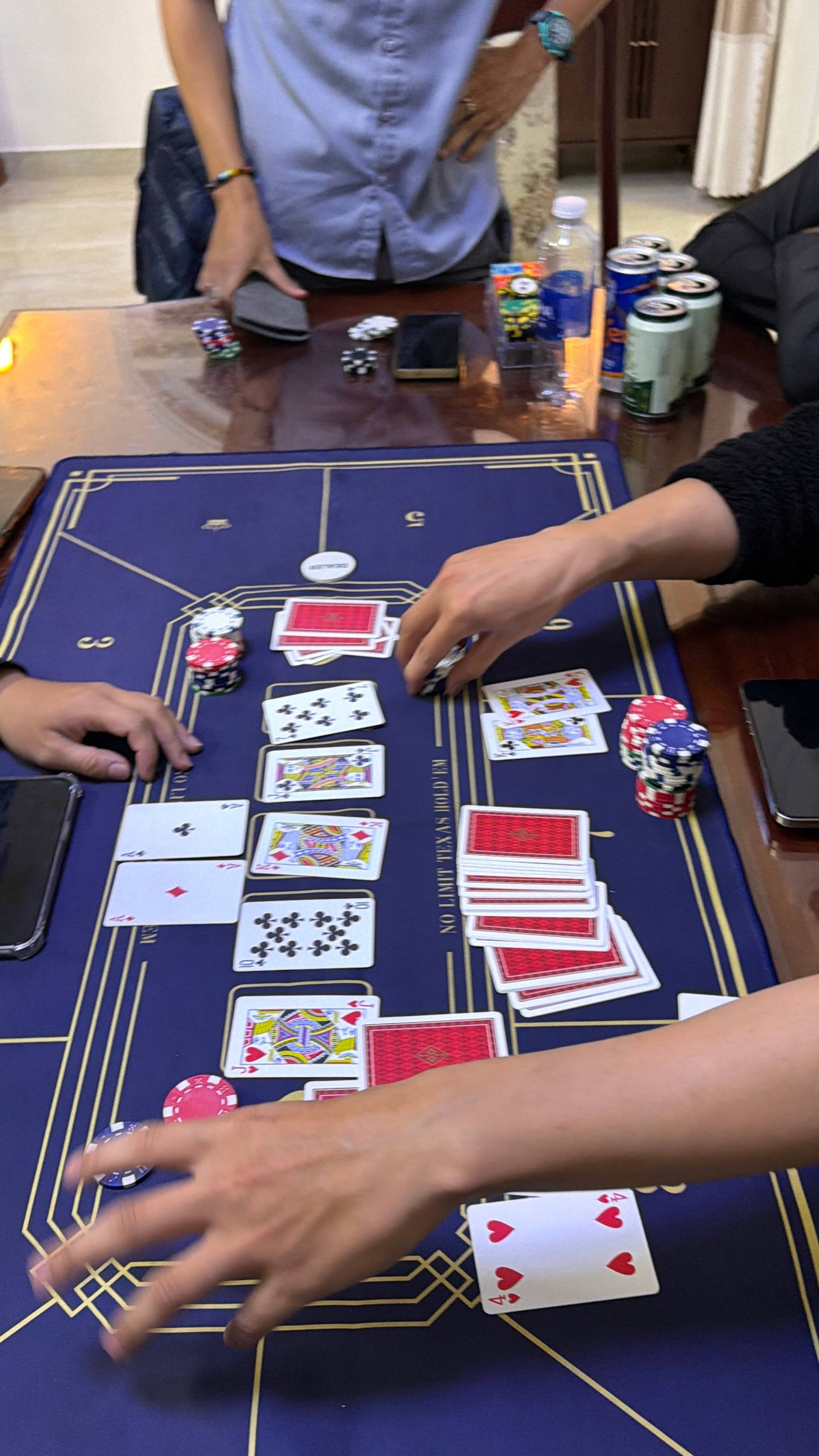 Two focused players engage in an intense baccarat game at a wooden table, with sunlight streaming through large windows illuminating the blue gaming mat and scattered poker chips. The intimate scene captures the concentration and strategy of card game play in a Vietnamese setting.