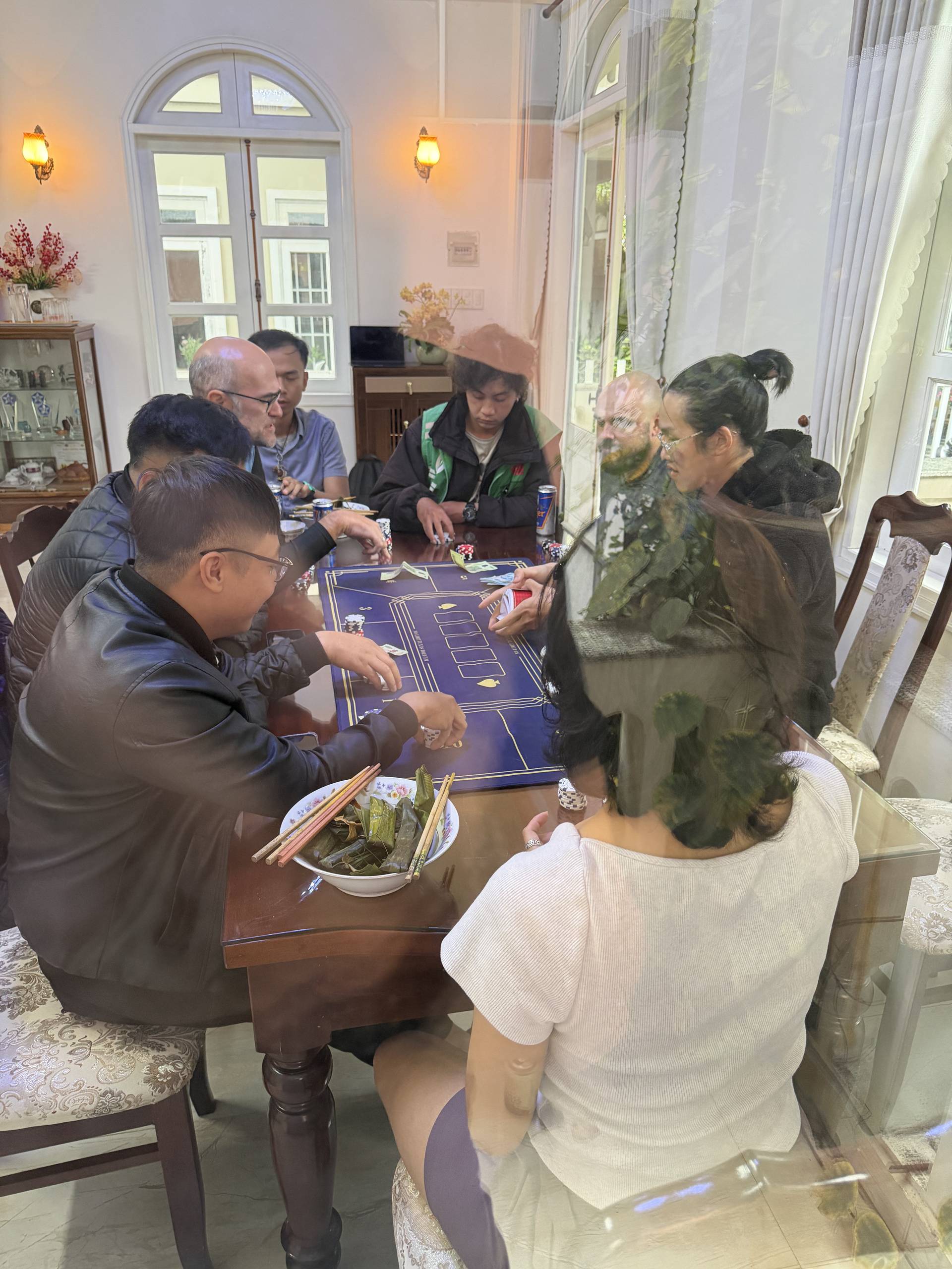 A mixed-age group gathers for baccarat in an elegantly appointed room with soaring white-framed windows and cream drapery. The intimate gaming experience showcases Vietnamese hospitality traditions, blending multiple generations in casual social entertainment.