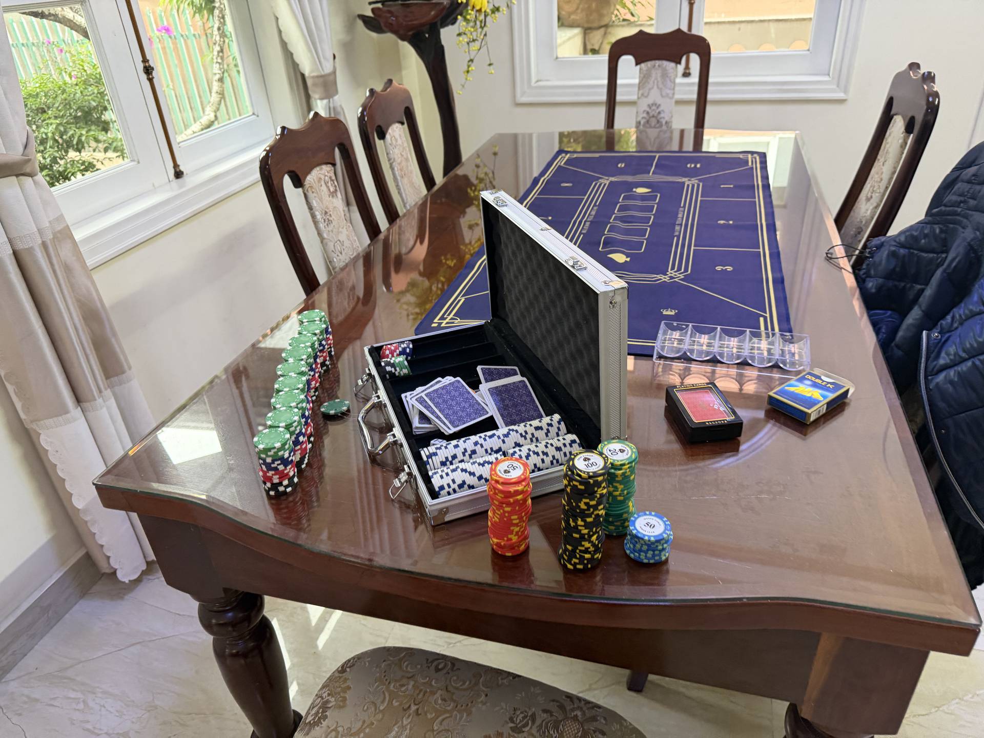 A professional baccarat gaming setup awaits players in a bright, elegantly furnished room with expansive windows overlooking lush gardens. Stacked poker chips and deck boxes are precisely arranged on the blue gaming mat, ready for an evening of entertainment.