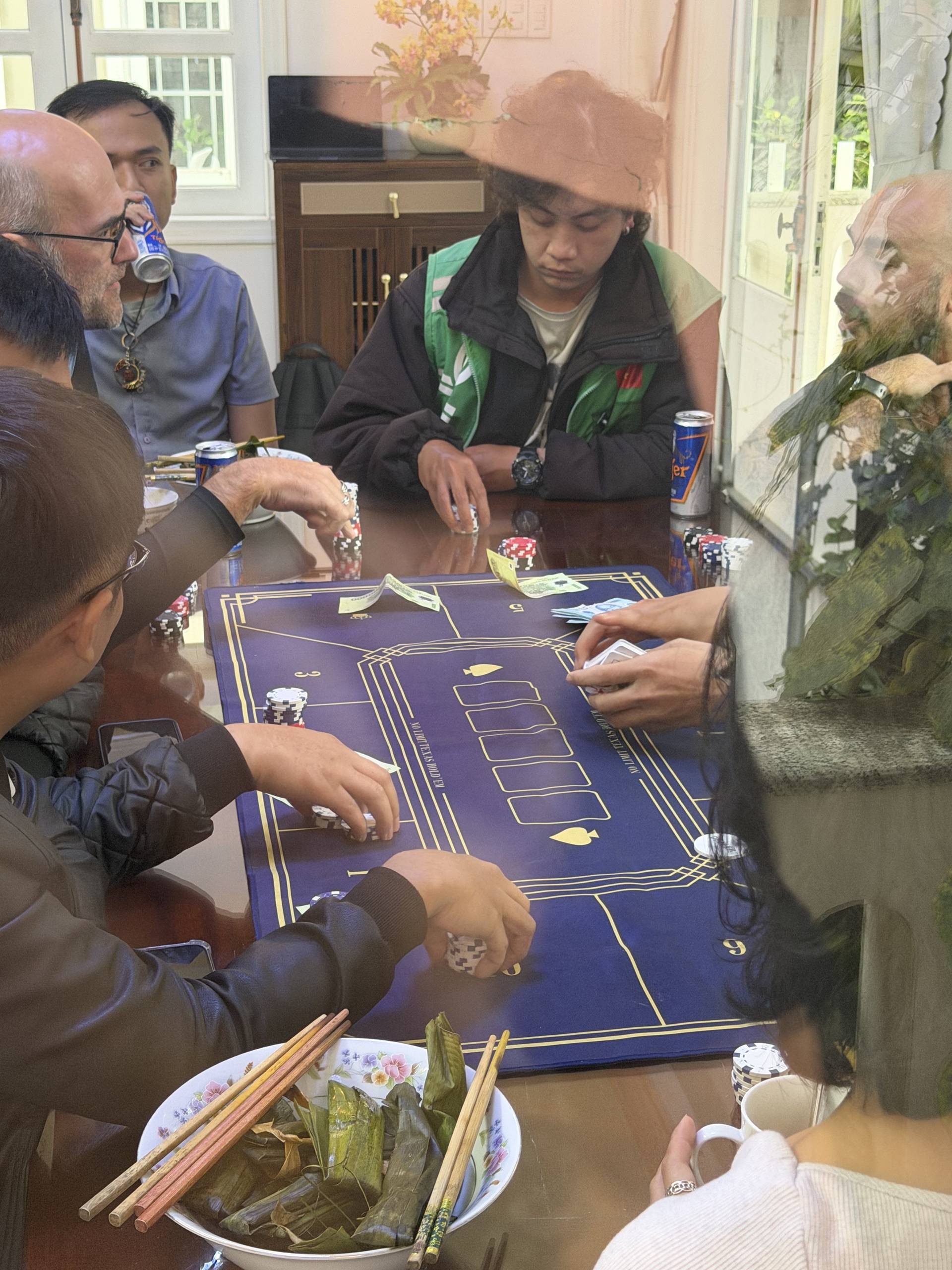 A diverse group plays baccarat around a wooden table in an elegant indoor setting with tall windows overlooking a garden. Vietnamese traditional snacks are served alongside the gaming experience, creating a blend of social entertainment and cultural hospitality.