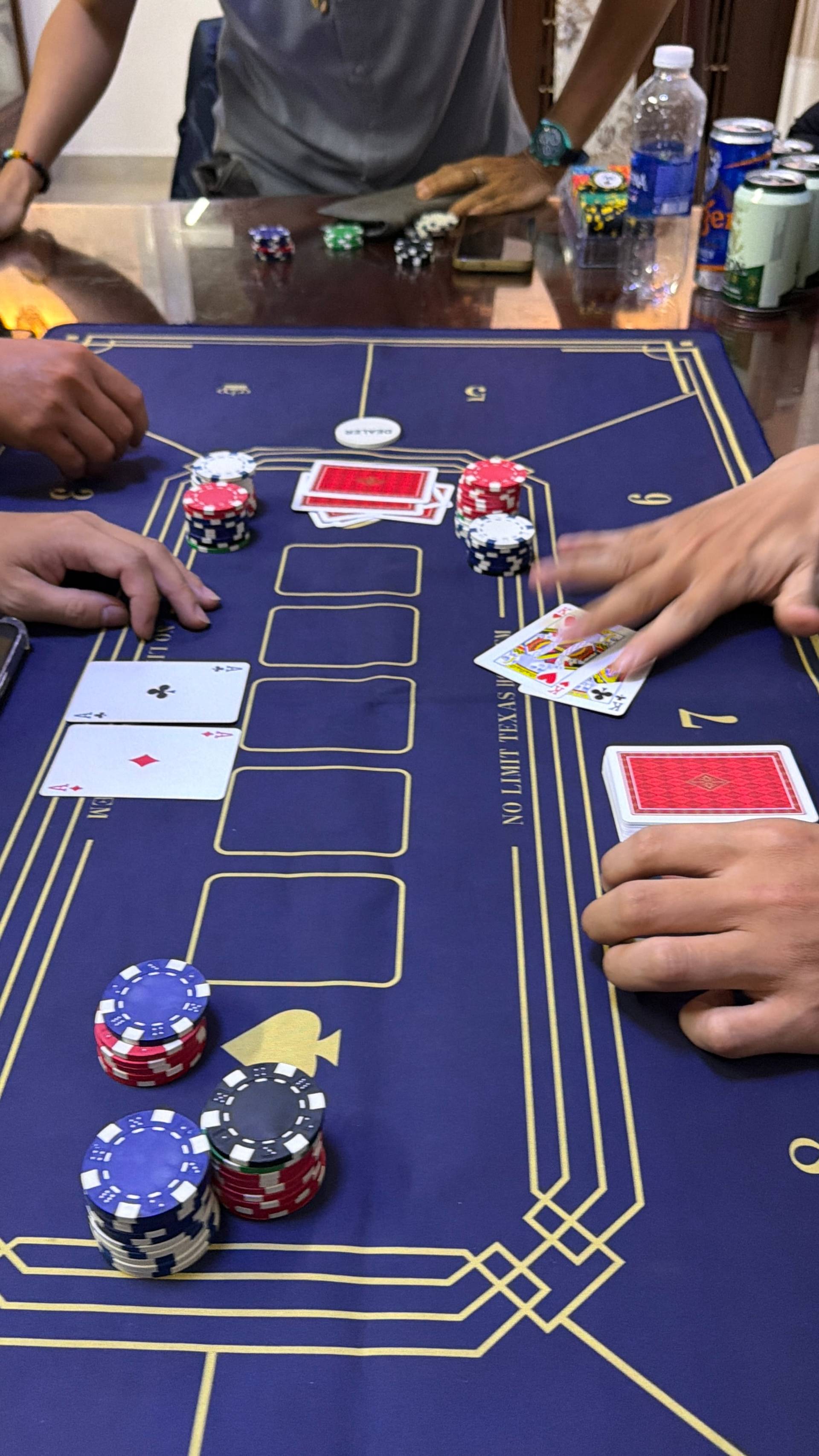 A detailed close-up of an active baccarat game showing dealt cards, stacked poker chips in red, white, and black, and hands positioned around a professional blue gaming mat. The scene captures the intensity and focus of the card game in a social setting.