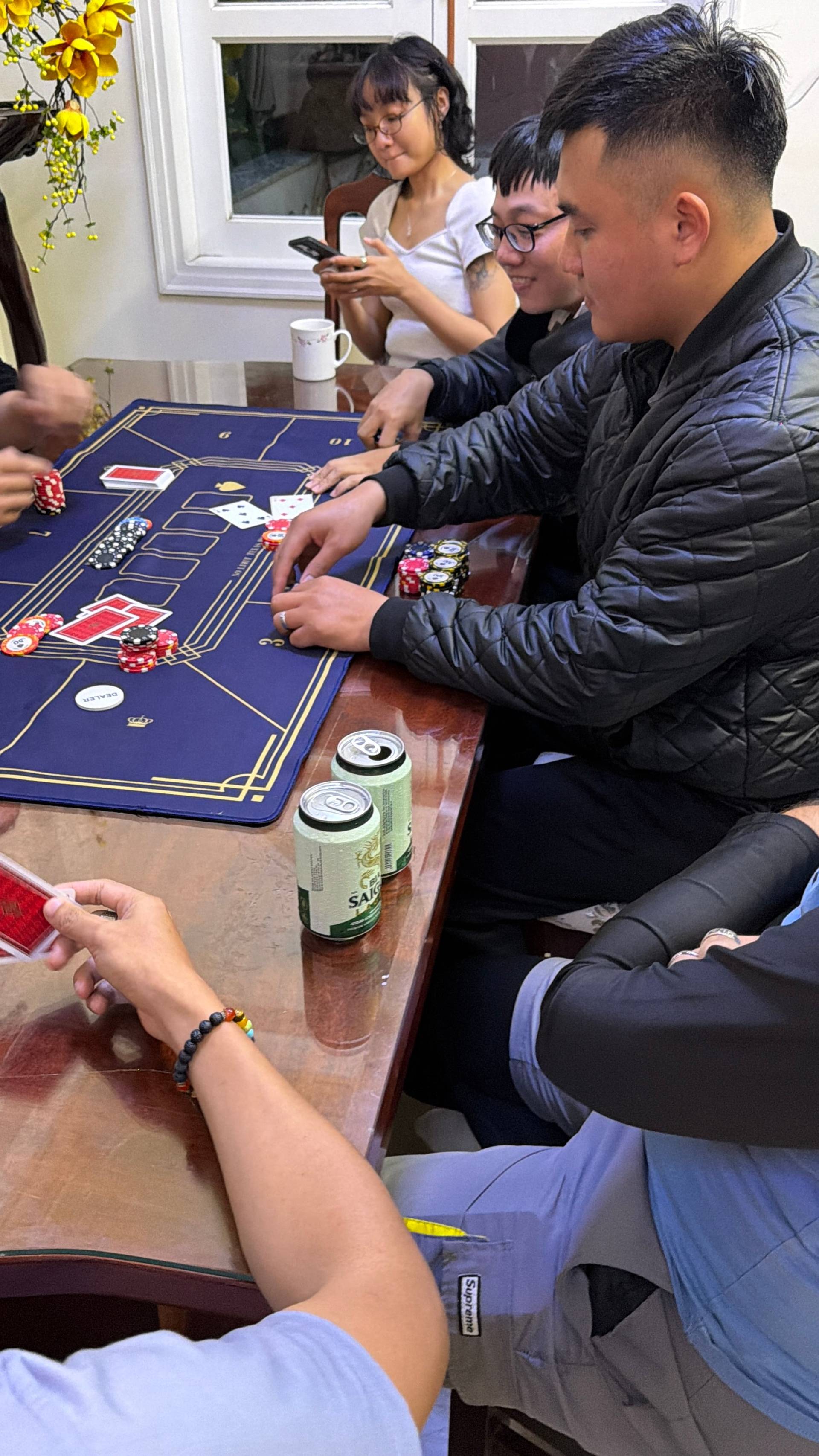 A group of friends gathered around a wooden table playing baccarat in a bright, airy indoor space with decorative plants and windows overlooking lush greenery. Colorful poker chips, playing cards, and refreshments create an atmosphere of casual social gaming in a cozy Đà Lạt home setting.