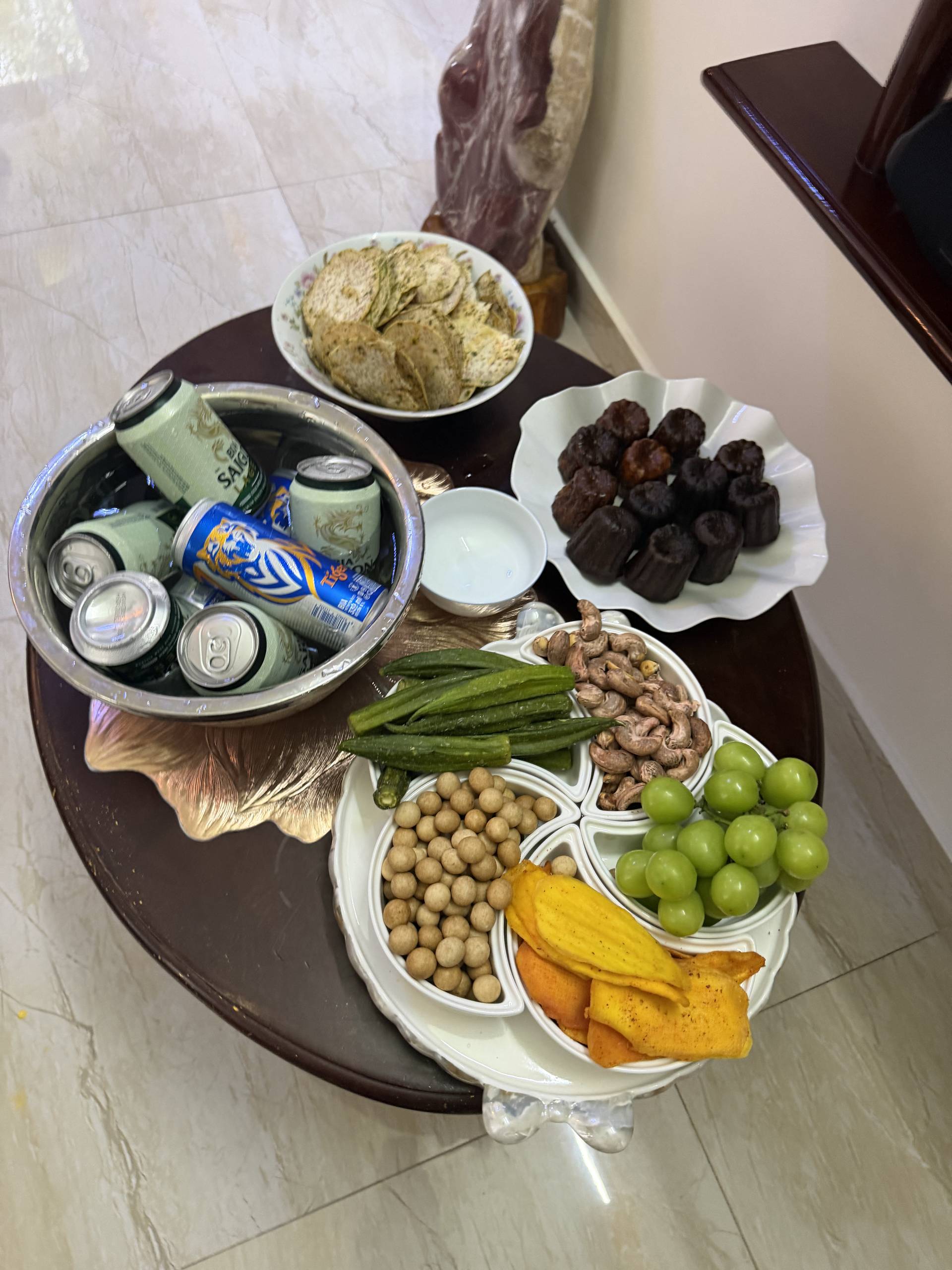 An overhead arrangement of Vietnamese party snacks and refreshments including grilled vegetables, nuts, chocolate confections, and beverages displayed on a decorative dark wooden table.