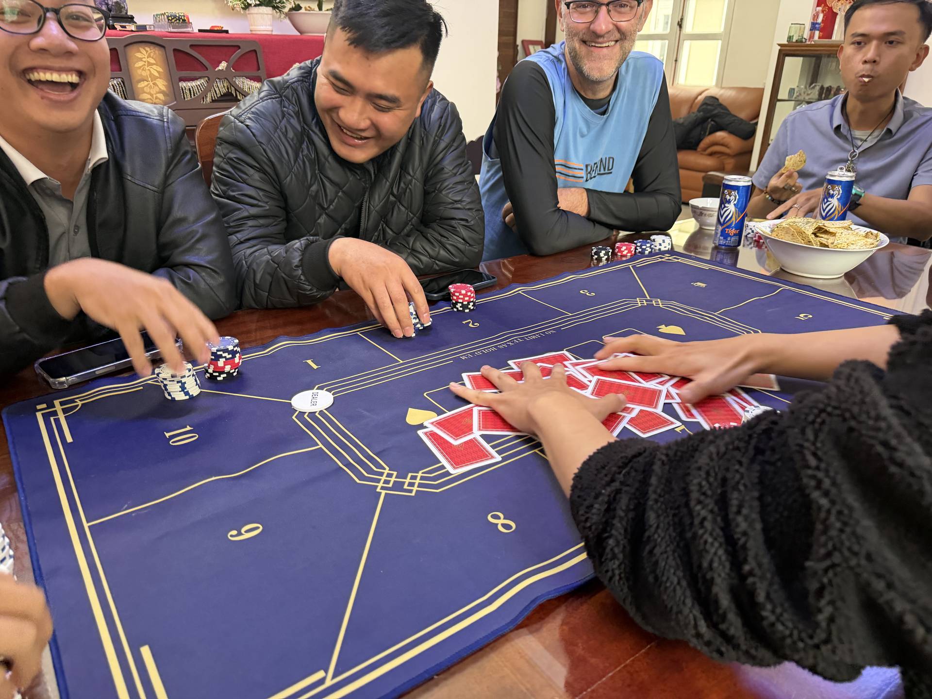 A lively group gathers around a professional baccarat table for an indoor gaming event in Đà Lạt, enjoying cards, chips, and refreshments in an elegant home setting.