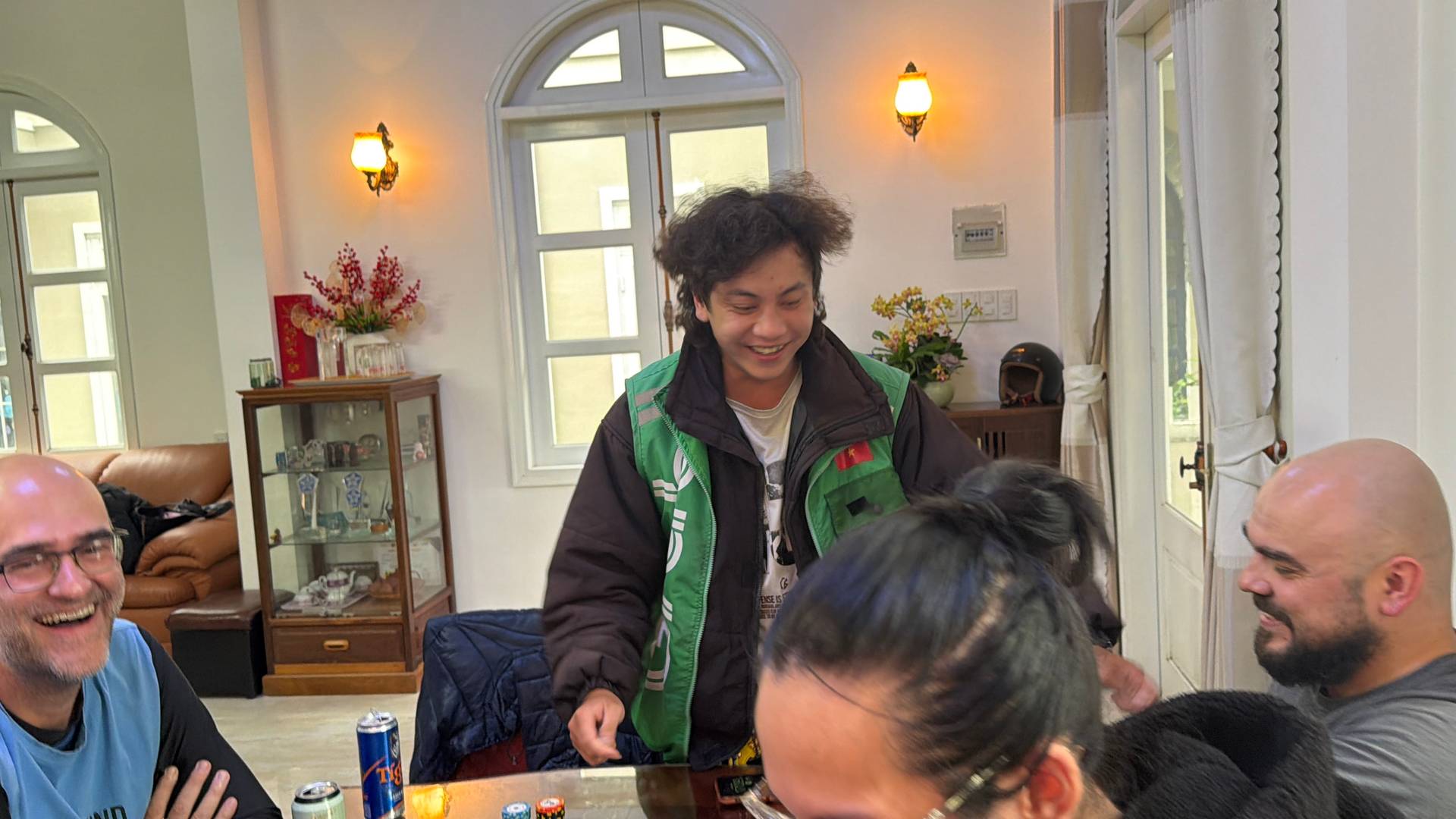 Close-up moment from a social gathering at an elegant Đà Lạt home, showing a group engaged in friendly conversation around a wooden table with refreshments.