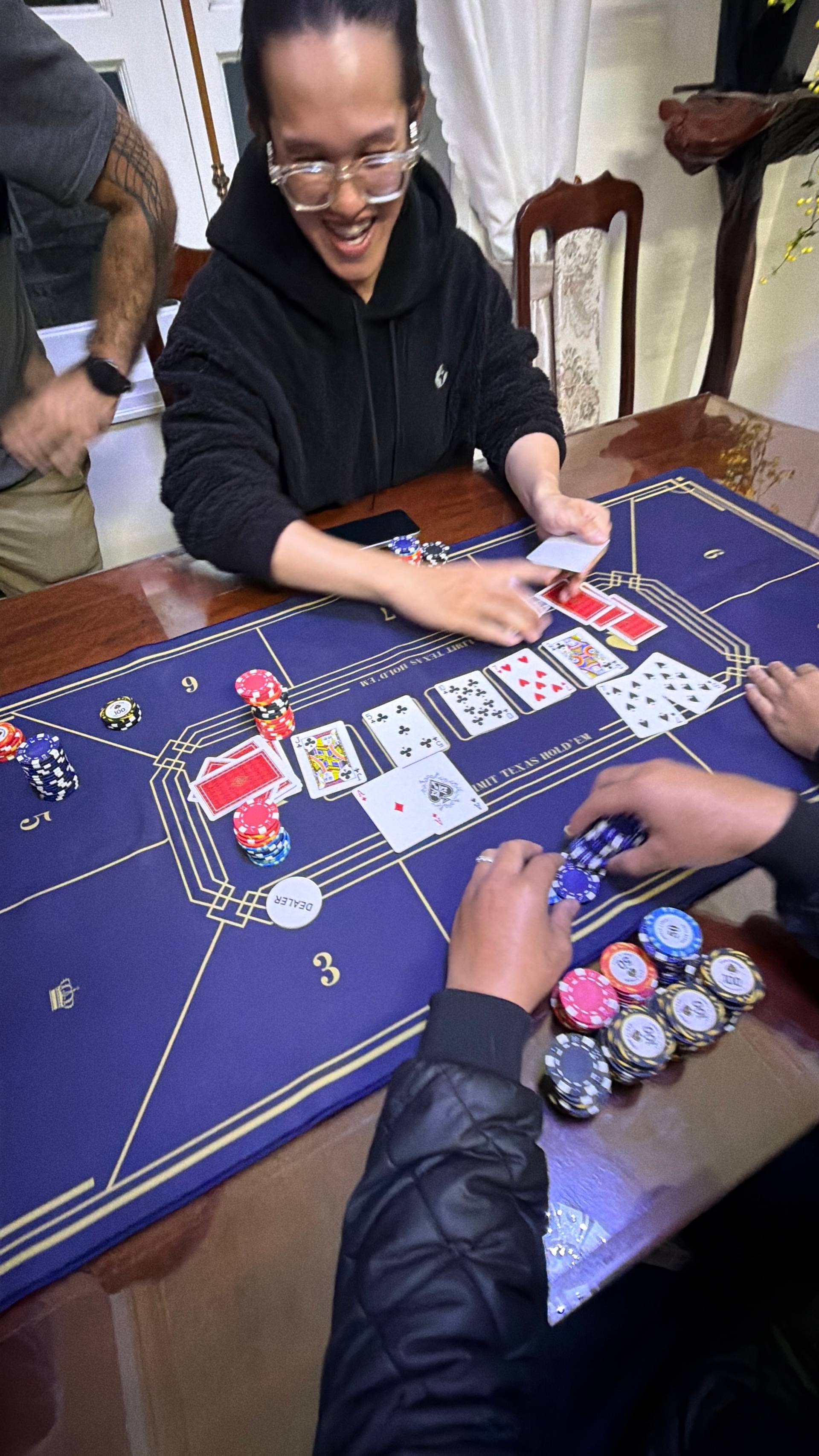 An enthusiastic player engages at the baccarat table during a Đà Lạt villa gaming night, with cards, chips, and a warm residential backdrop creating an intimate event atmosphere.