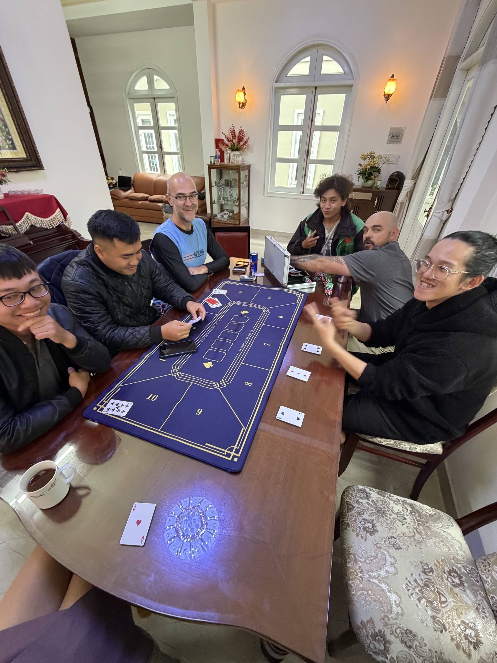 A lively group gathering around a baccarat table in a spacious Đà Lạt villa living room, with participants enjoying cards, chips, and drinks in an elegant residential setting.