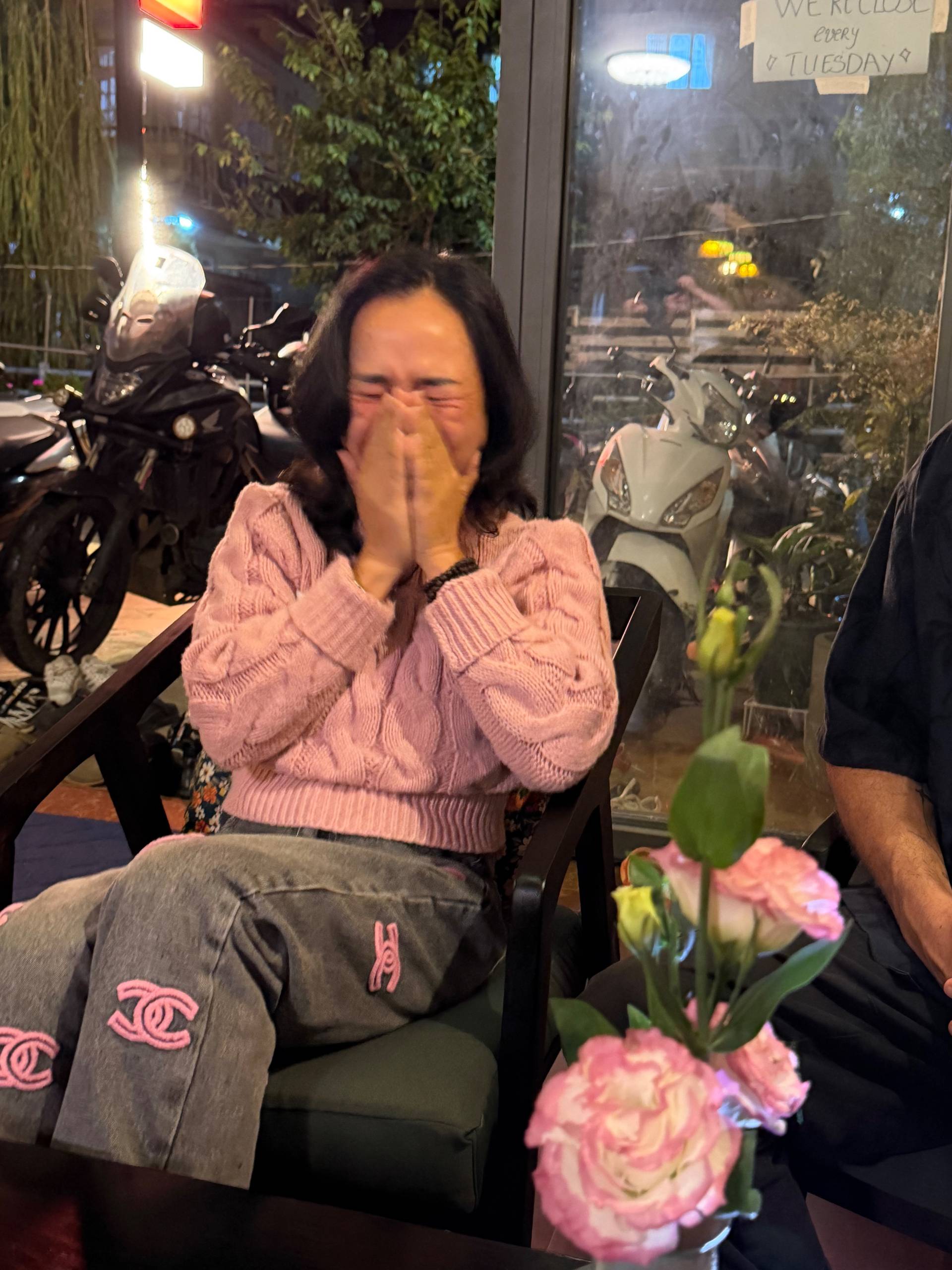 A cozy café scene in Đà Lạt with a woman laughing heartily while sitting by a window overlooking parked motorcycles and street lights at night.