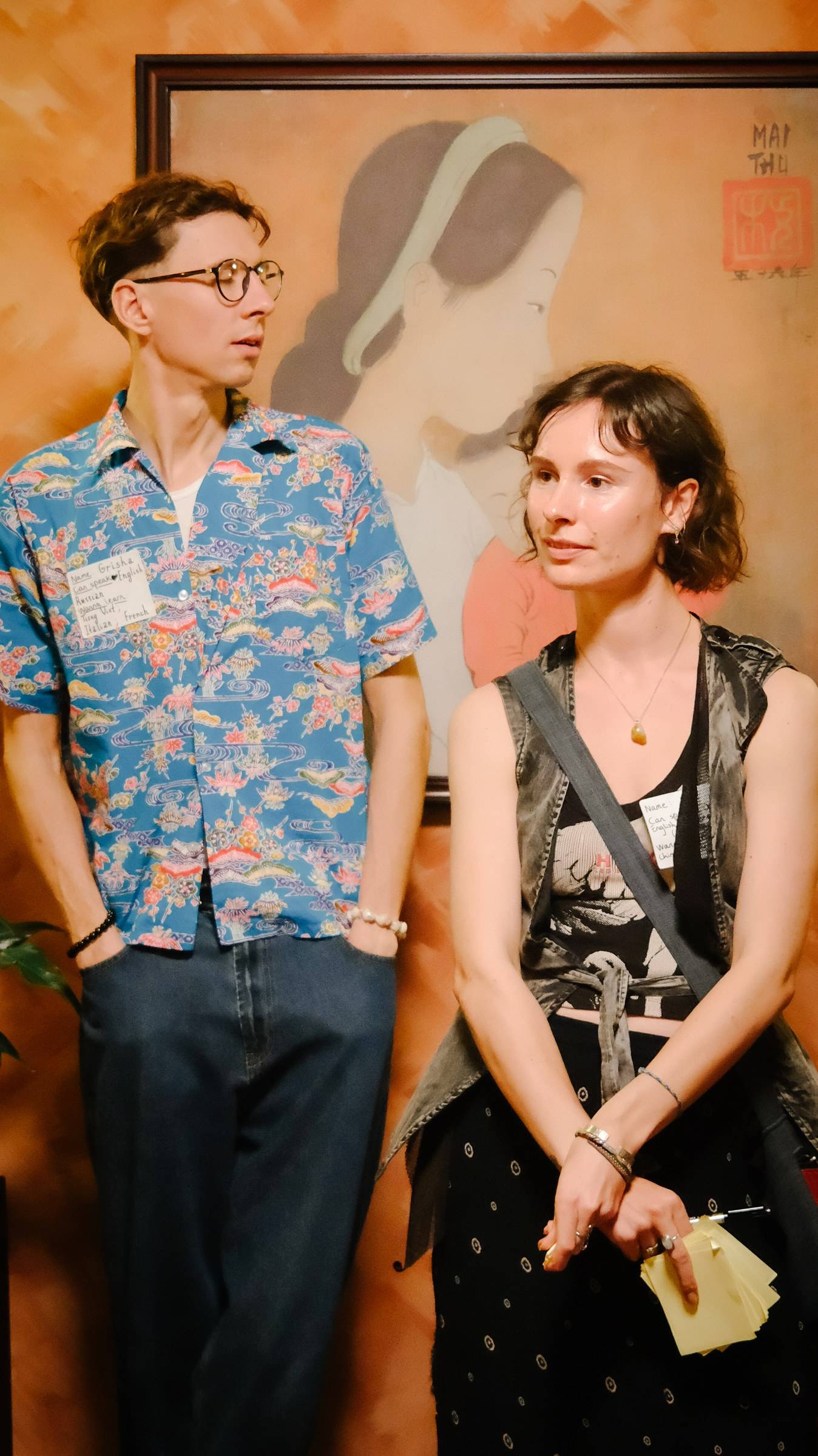 Two individuals stand in front of a traditional Vietnamese painting, engaging in conversation with a relaxed and casual demeanor.