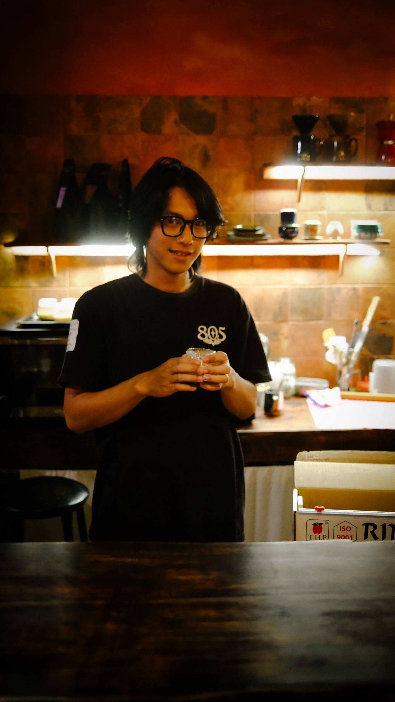 A person in a café setting in Đà Lạt, Vietnam, holding a drink and wearing a black t-shirt with '805' printed on it.