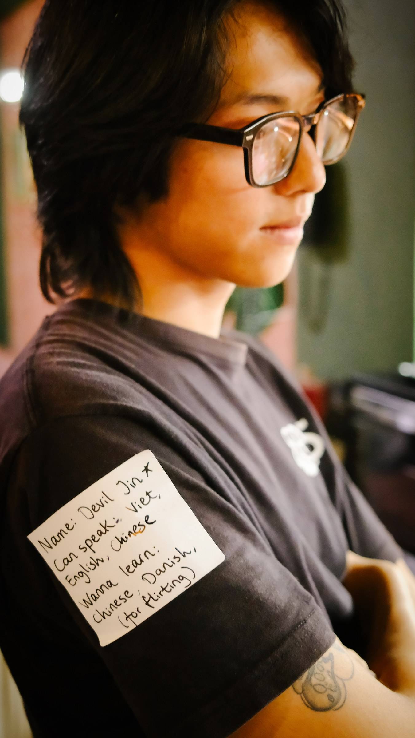 A person wearing a black t-shirt with handwritten notes about language skills and interests is seen in what appears to be an indoor setting in Đà Lạt, Vietnam.