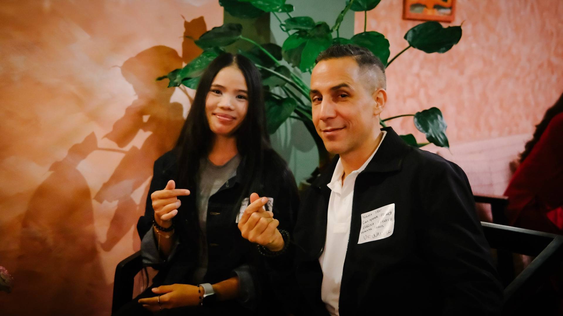 Two individuals smile warmly in a cozy indoor setting with vibrant wall art and lush greenery, suggesting a relaxed social gathering or event in Đà Lạt.