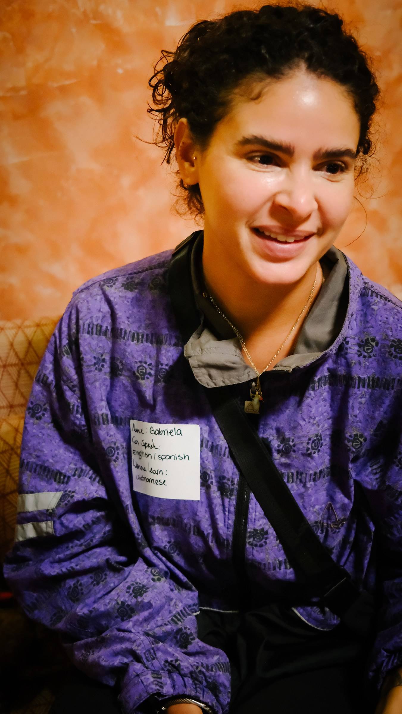 A person wearing a purple jacket with a name tag that reads 'Gabriela' and indicates she can speak English/Spanish and wants to learn Vietnamese at an event in Đà Lạt, Vietnam.