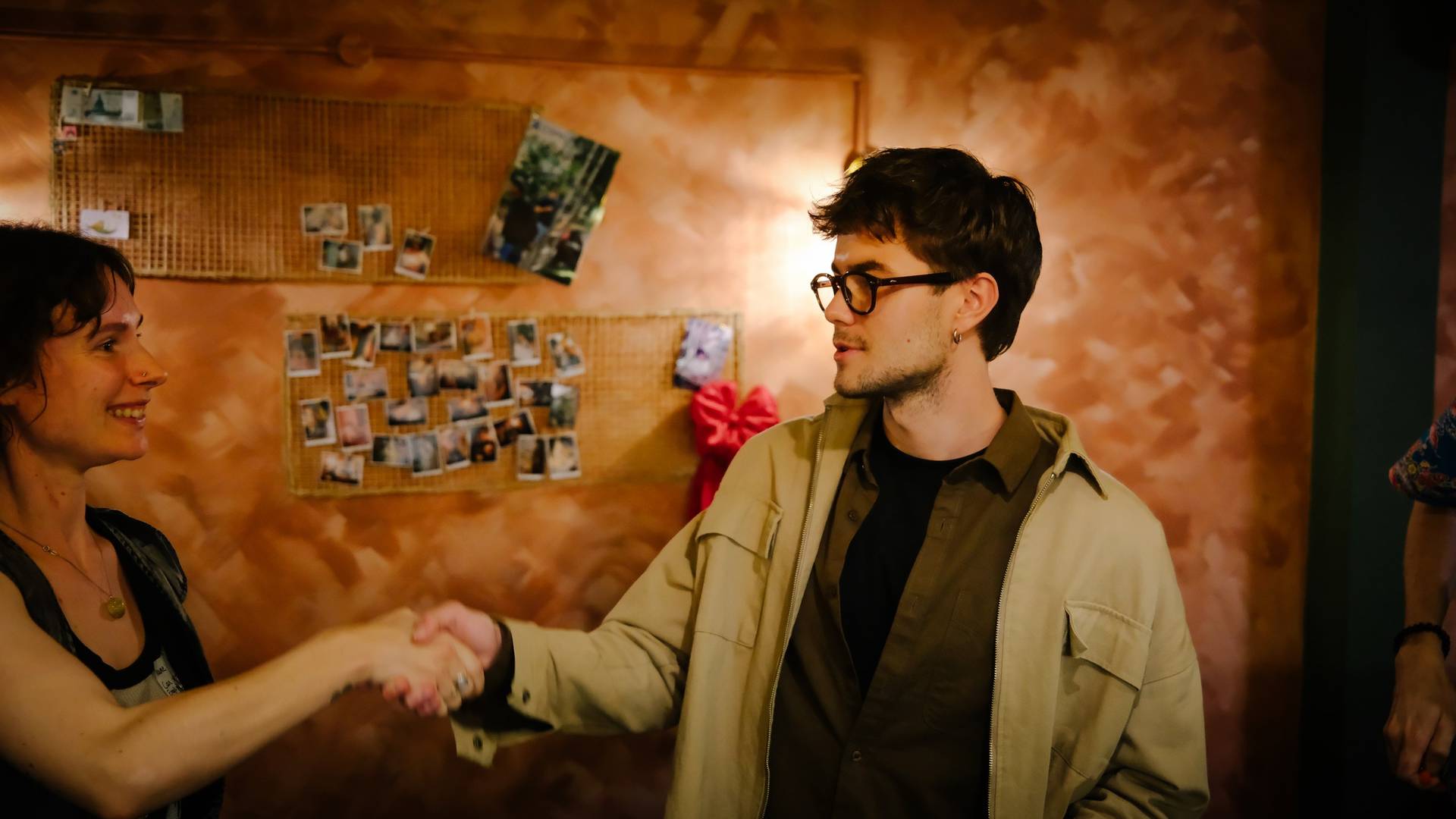 Two individuals share a warm handshake in a cozy, rustic setting adorned with personal photographs and vibrant decor, reflecting a sense of camaraderie and connection.