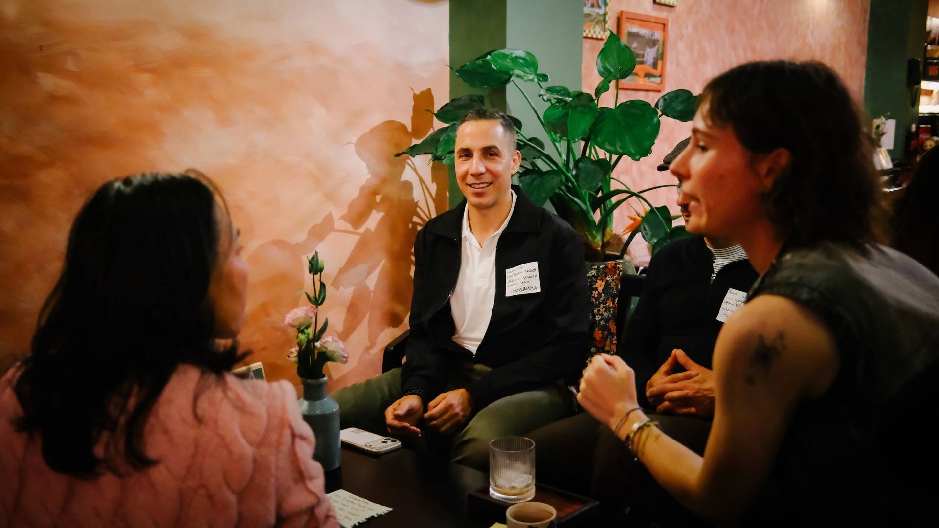 A group of people engaged in conversation over drinks in a cozy café setting with warm lighting and decorative plants, reflecting a relaxed and intimate atmosphere.