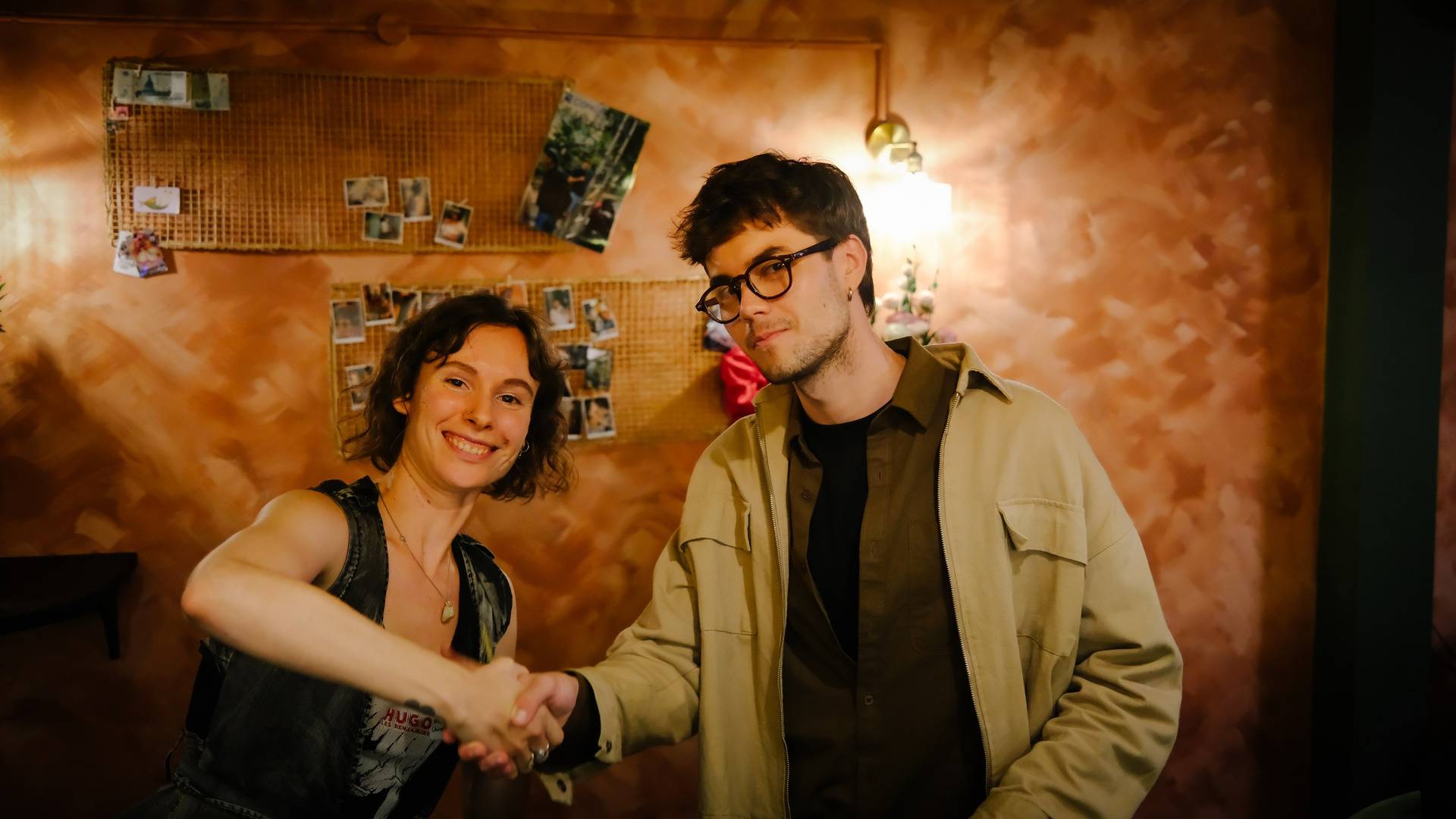 Two individuals share a warm handshake in a cozy indoor setting with personal photos and decor, suggesting a friendly or celebratory gathering.