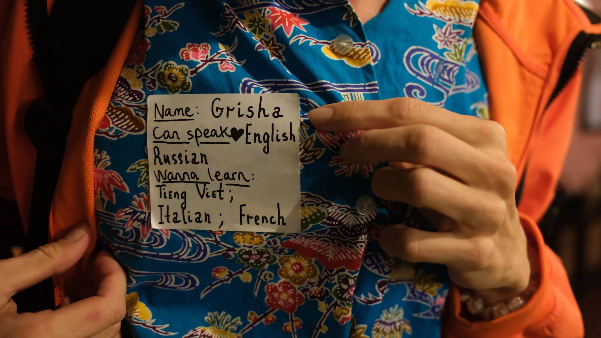 A person in Đà Lạt, Vietnam, wearing a colorful shirt with a handwritten note pinned to it, indicating their language skills and interest in learning Vietnamese.